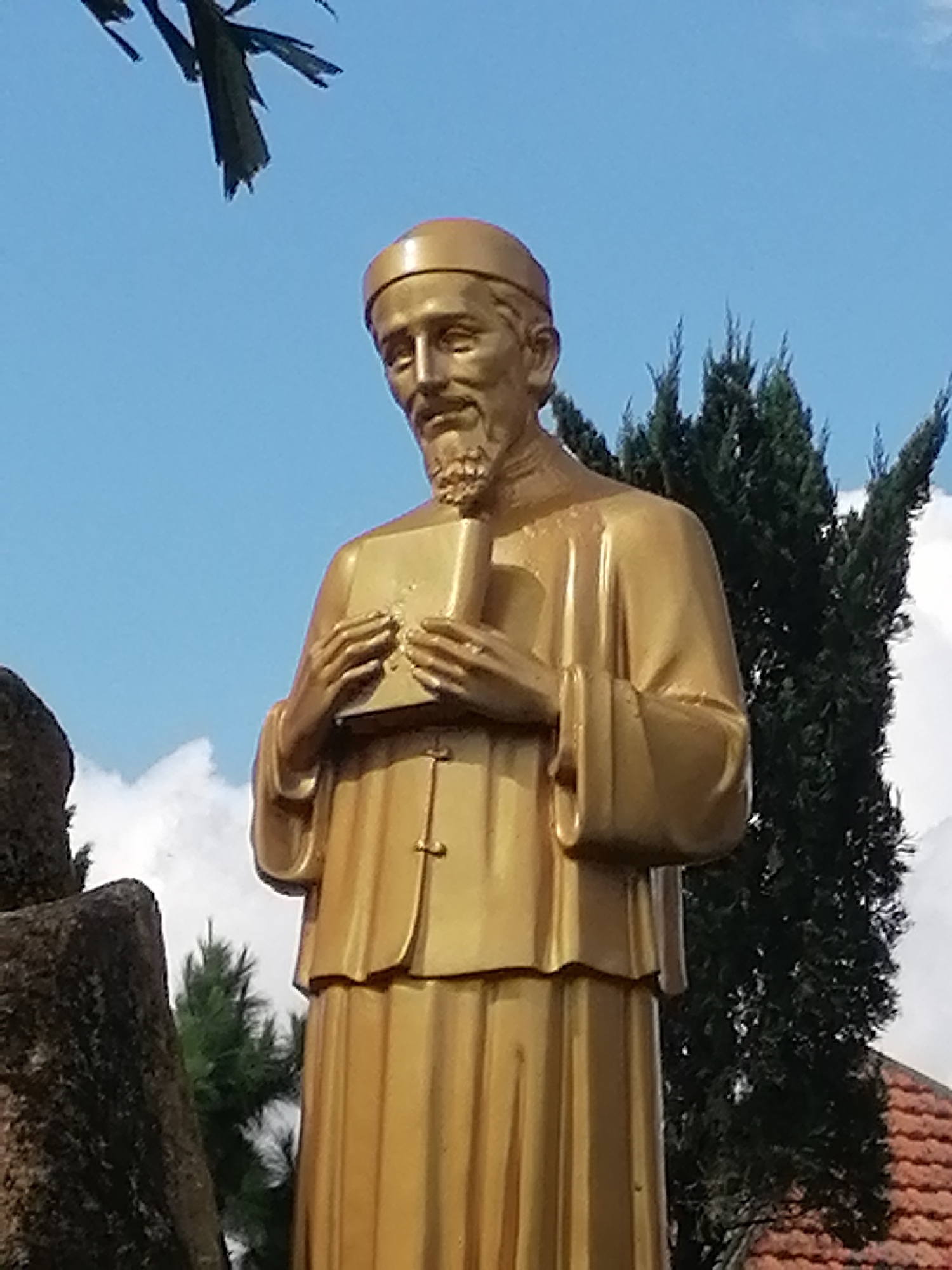 A golden statue of a man wearing a cap and robes stands in front of a backdrop of blue sky with scattered clouds and a tall evergreen tree. The statue's hands are clasped around a book. A broken tree branch is visible in the upper left corner of the image. The statue is located in front of a building with a red tile roof. The statue is believed to be of Saint John Baptist Mary Vianney, located at the Saint John Baptist Mary Vianney Church in Da Lat, Vietnam.