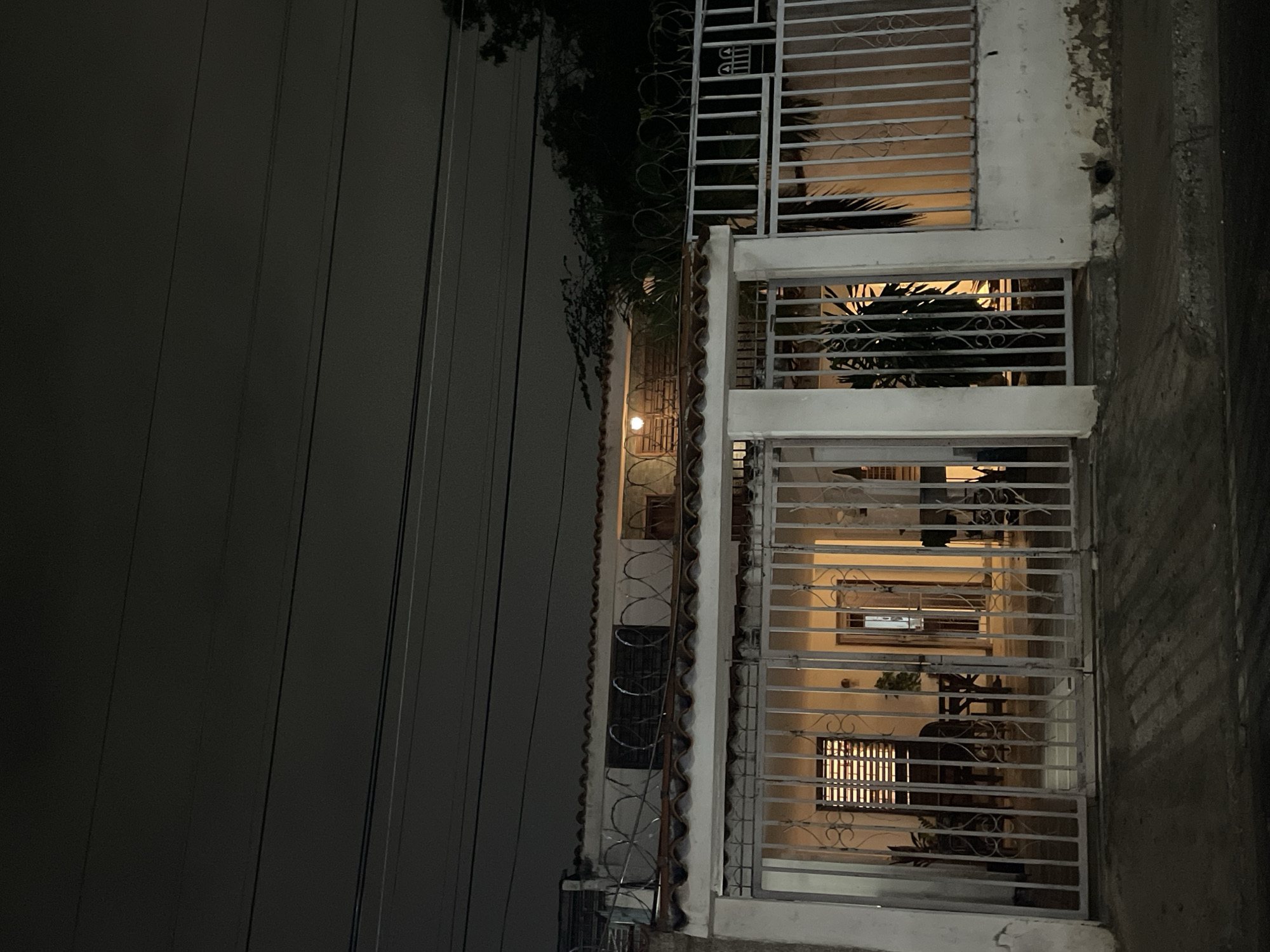 The image depicts a building exterior at night in Unknown, Unknown. The building has at least three floors, each with a window covered by metal bars. There is a light fixture visible on the facade of the building. Above the building, there are trees and power lines. The building's exterior is weathered and shows signs of age.