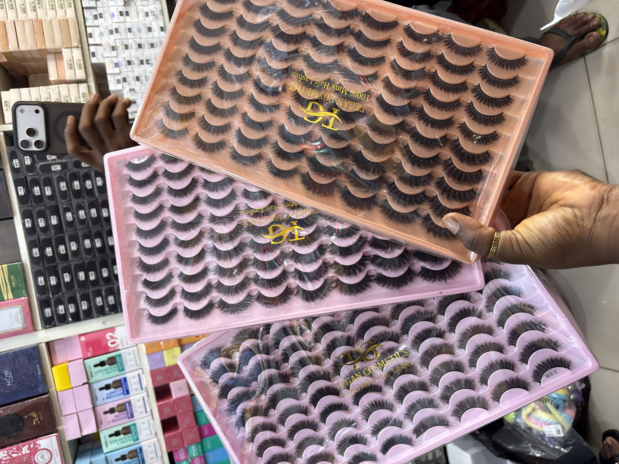 The image displays a close-up view of three pink-colored trays filled with artificial eyelashes. Two of the trays are in the foreground, tilted at an angle, showcasing multiple rows of eyelashes. The leftmost tray has the text "MIX-5" visible on one of its compartments. The middle tray features a gold-colored logo and text that reads "TEGAN COSMETICS 100% Mink Hair Lashes". The third tray, partially visible to the right, also contains similar artificial eyelashes. In the background, shelves are stocked with various cosmetic products, including boxes of serums and other beauty items. A person's hands are visible holding the trays, with a ring on the right hand. The reflection of a smartphone, held by the person, is visible in a mirror or shiny surface in the background, showing a partial view of the user. The overall setting appears to be a retail environment, likely a cosmetics store. The presence of the "TEGAN COSMETICS" branding and the assortment of beauty products suggest this could be a retail location within Jos, Nigeria, a city known for its commercial activity, including the sale of beauty products.