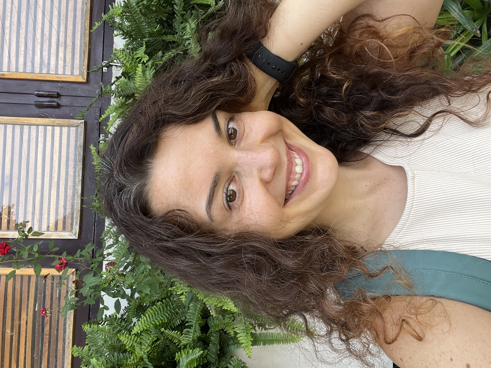 A woman with long, curly brown hair and gray streaks smiles at the camera, her right hand resting on the back of her head. She wears a white ribbed tank top and a teal backpack strap is visible over her left shoulder. She has brown eyes and appears to be outdoors, with lush green ferns and a wooden structure with striped windows in the background. The wooden structure resembles shutters or window coverings, and a climbing vine with red flowers is present. The environment suggests a tropical or subtropical location. The image appears to be taken in Mũi Né, Vietnam, given the architectural style and vegetation.