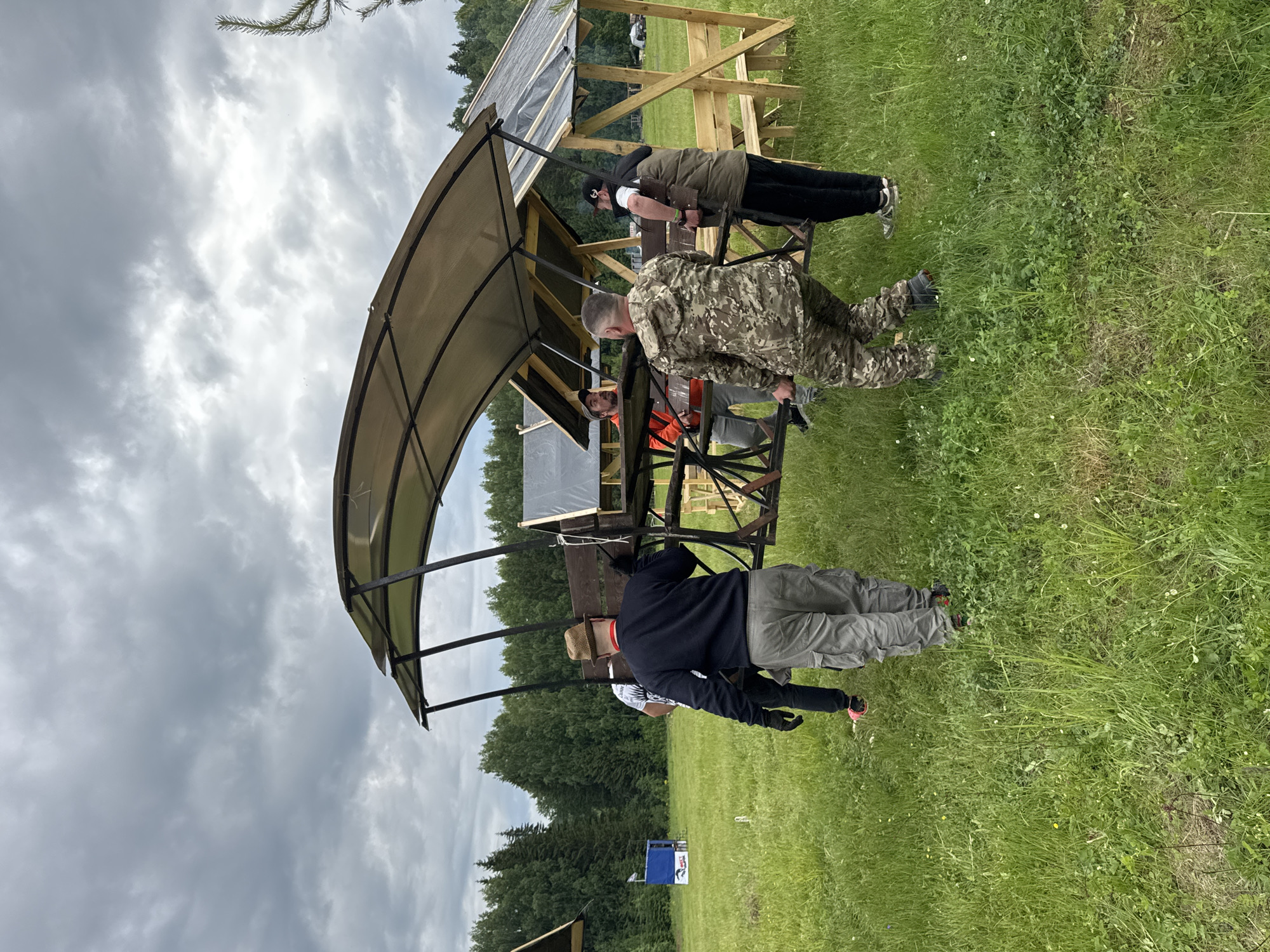 This outdoor scene, set in Большая Кивара, Russia, captures several men engaged in an activity under an overcast sky. The setting is a large grassy field with varying shades of green, dotted with clover and other vegetation, extending to a horizon lined with dense green trees. The sky above is largely cloudy, with patches of brighter, high-altitude clouds suggesting diffused daylight.
In the foreground and mid-ground, a prominent structure with a curved, light-colored canopy supported by dark metal poles dominates the left side of the frame. Behind it, a wooden frame structure, partly covered with a black tarp, is visible. Several men are gathered around these structures.
One man in the immediate foreground, partially facing away from the viewer, wears a dark hoodie, light grey cargo pants, black gloves, and a light-colored hat. To his right, another man, dressed in camouflage fatigues, is leaning forward, seemingly focused on something at a lower level. Further back, a man in an orange vest is partially visible, and another man in a dark jacket, black pants, and a baseball cap stands near the wooden frame.
In the distant background, a blue and white object, possibly a cooler or container, shows the partial text "RUS" or "РК". The overall impression is of an organized outdoor event, perhaps a training exercise, a sporting competition, or a recreational activity, taking place on a cloudy day. The men appear focused, but their emotions are not clearly discernible.
