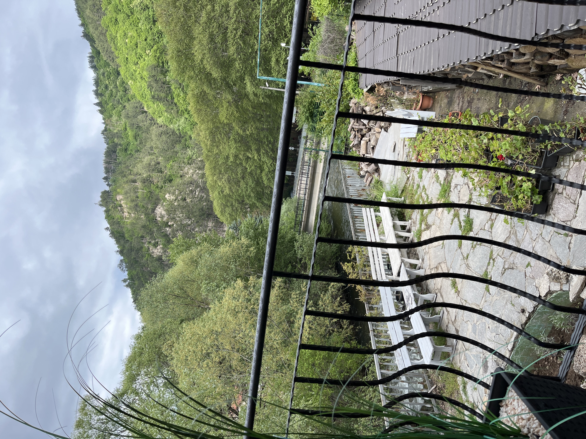 View from a wrought iron balcony in Bachkovo, Bulgaria, overlooking a stone-paved patio with white benches, potted plants, and stacked firewood. Beyond the patio, a bridge spans a river, leading to a tree-covered mountain. The sky is overcast.