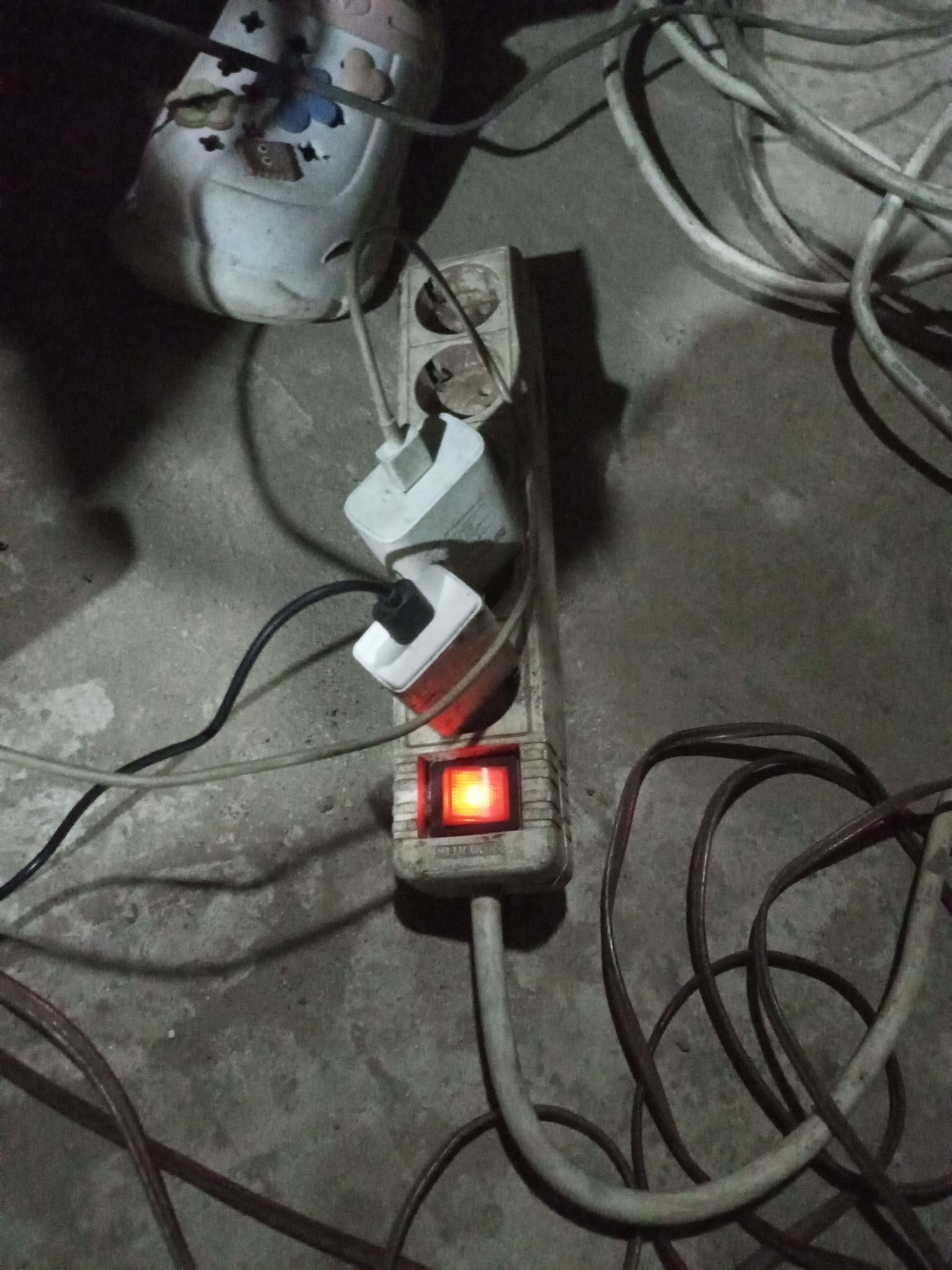 An overhead, medium shot captures a power strip plugged into a wall outlet, lying on a concrete floor. The power strip is off-white and rectangular, with two visible sockets. A white electrical adapter is plugged into the top socket, and another white adapter with a red trim is plugged into the bottom socket. A black power cord extends from the bottom adapter and snakes across the floor. A single red illuminated switch is visible on the front of the power strip. A light-colored shoe with blue, black, and light brown floral patterns rests on top of the power strip, partially obscuring it. Several electrical cords, including a thick grey one and a thinner brown one, are coiled and spread across the concrete floor. The scene is dimly lit, casting shadows on the ground. The location is identified as Unknown, Unknown.