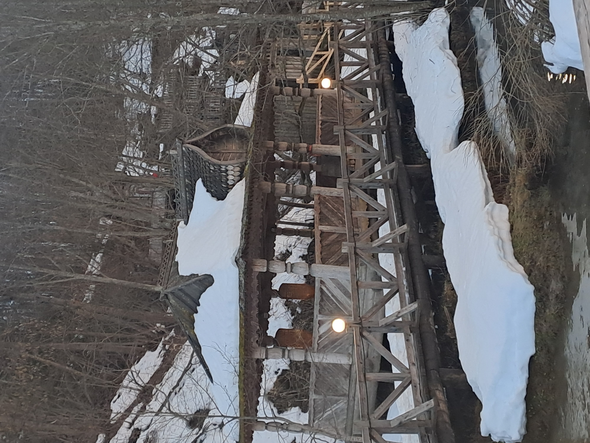 The image depicts a snow-covered scene in Vzglyadnevo, Russia. There is a wooden structure, possibly a bridge or walkway, with wooden supports and two visible lights along its length. Adjacent to the structure is snow on the ground and on the roofs of buildings, some with intricate tilework. Trees, mostly bare of leaves, are present throughout the scene. The overall environment suggests a cold, possibly winter, setting.