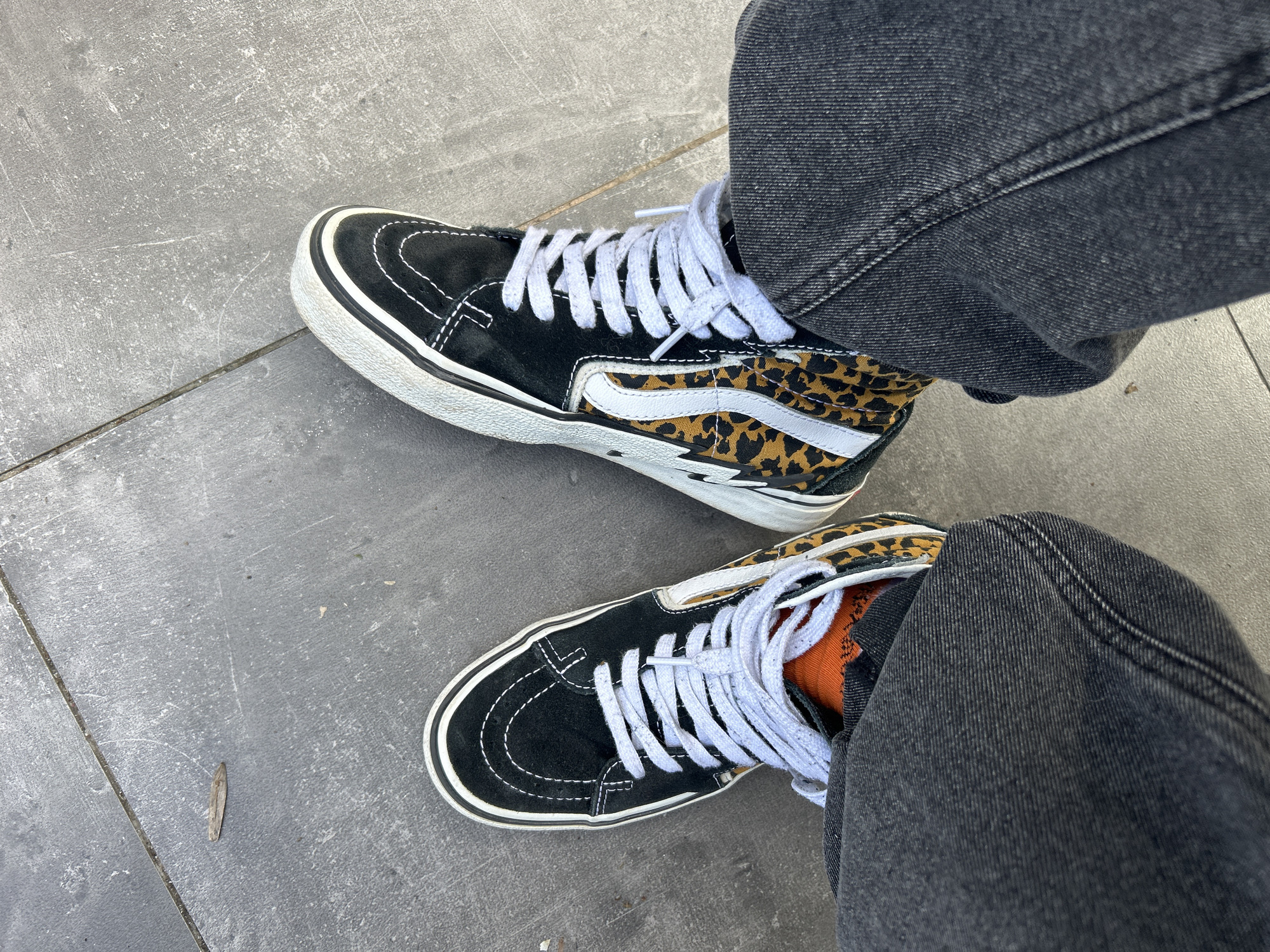 A person is standing on a gray tiled surface, likely a sidewalk. They are wearing black and leopard print high-top Vans sneakers with white laces and dark gray jeans. An orange sock is visible on their right foot. A small, dry leaf is present on the tiled surface. The location is unspecified and cannot be confirmed as Sofia, Bulgaria based solely on the image.