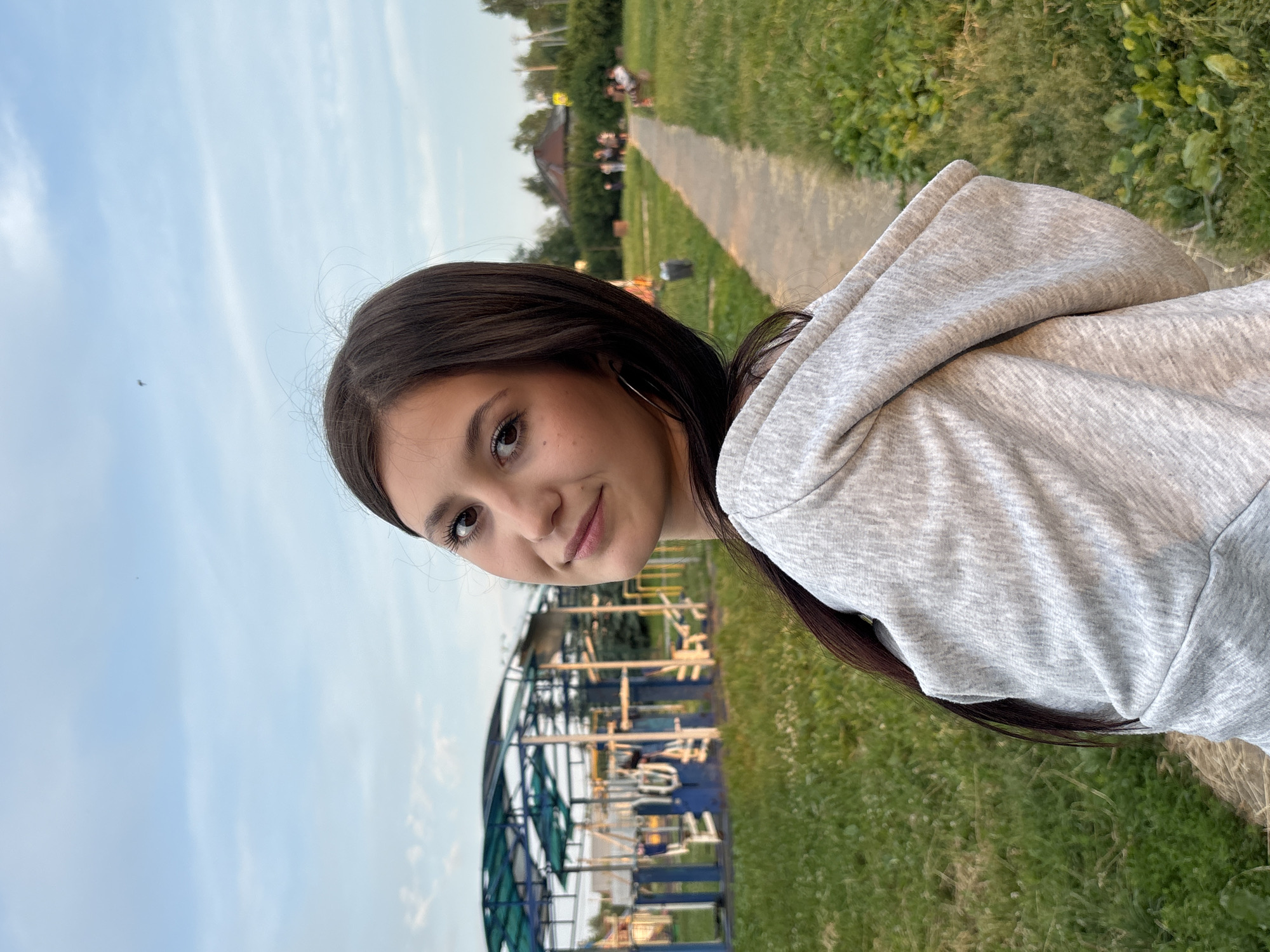 A young woman with dark hair is captured in a close-up, profile view, looking over her right shoulder towards the camera. She is wearing a light gray hooded sweatshirt. Her expression is a slight smile. Behind her, a playground structure with blue and green elements is visible, along with trees and a pathway in the background. The setting appears to be an outdoor park or recreational area in the city of Фоки, Russia. The lighting suggests it is late afternoon or early evening.