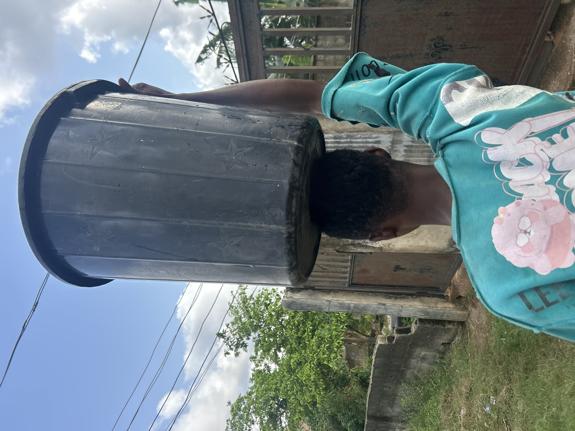 A person is depicted carrying a large, black plastic bucket on their head in an outdoor setting. The image is oriented sideways. The person's head, with short, dark, curly hair, is visible beneath the bucket, and their right arm is raised, possibly to steady the load. They are wearing a long-sleeved teal top with white and pink graphic designs. Some stylized text and imagery, including shapes that resemble bones or cartoon elements, are visible on the sleeve and front of the top.
The black bucket has several embossed five-pointed stars on its textured surface. In the background, a bright blue sky with fluffy white clouds is visible. Utility lines cross the sky. Below the horizon, there are green trees with dense foliage, concrete walls, and what appears to be a dark metal gate or door in a concrete structure. The ground beneath the subject has patches of grass and dirt. The scene takes place during the daytime under clear weather conditions.