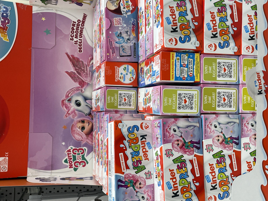 The image displays a retail store shelf in Monsummano Terme, Italy, featuring a tiered arrangement of Kinder Surprise and Kinder Applaydu products. The primary theme is unicorns and a female character with pink hair. A large cardboard header sign at the top showcases a pink-haired character looking at a white winged unicorn, with text in Italian reading "NOVITÀ 3 SU 3" (New 3 out of 3) and "SCOPRI IL MONDO DEGLI UNICORNI!" (Discover the world of unicorns!). Below this, multiple rows of Kinder Surprise boxes are stacked. White and red boxes labeled "Kinder Sorpresa" and "Senza Glutine" (gluten-free) are visible, some indicating "x3" for three eggs. Smaller individual boxes in pink, purple, and green are interspersed, many featuring "SCOPRI COME RICICLARE!" (Discover how to recycle!) with QR codes and "recycle info". Other boxes display text like "Scansiona la sorpresa nel mondo digitale" (Scan the surprise in the digital world) with an image of a phone showing a digital unicorn, and "Vinci fantastici premi!" (Win fantastic prizes!). The products are uniformly aligned, indicating a structured commercial display.