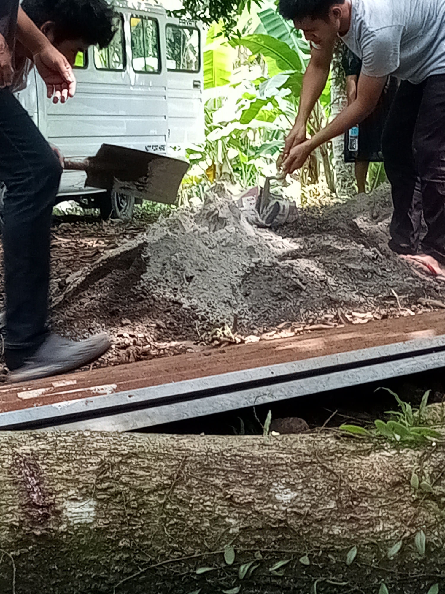 The image depicts two individuals, possibly men, handling a pile of gray material, likely soil or cement mix, on what appears to be a metal platform or the back of a vehicle. One person is holding a shovel, while the other is using their hands. A white vehicle is visible in the background, possibly a van or truck. The scene is outdoors, with trees and other vegetation present. A large log or tree trunk is in the foreground. I cannot verify the image is located in Davao City, Philippines.