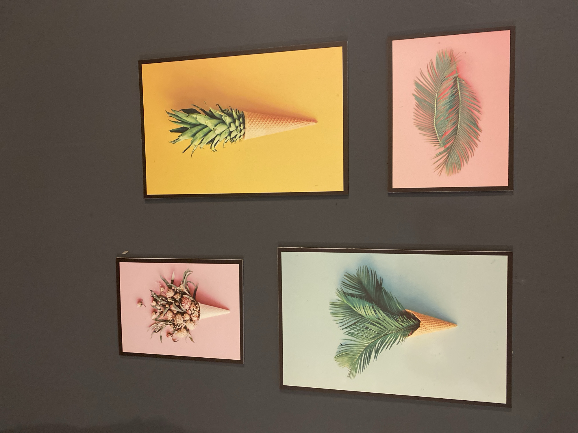 The image displays four framed prints on a dark gray wall. The prints are arranged in a two-by-two grid. The top left print features a bouquet of dried flowers in a waffle cone on a pink background. The top right print shows a pineapple in a waffle cone against a vibrant yellow background. The bottom left print depicts palm fronds emerging from a waffle cone on a light blue-green background. The bottom right print presents two palm fronds on a coral pink background. The location is a wall in Đà Nẵng, Vietnam.