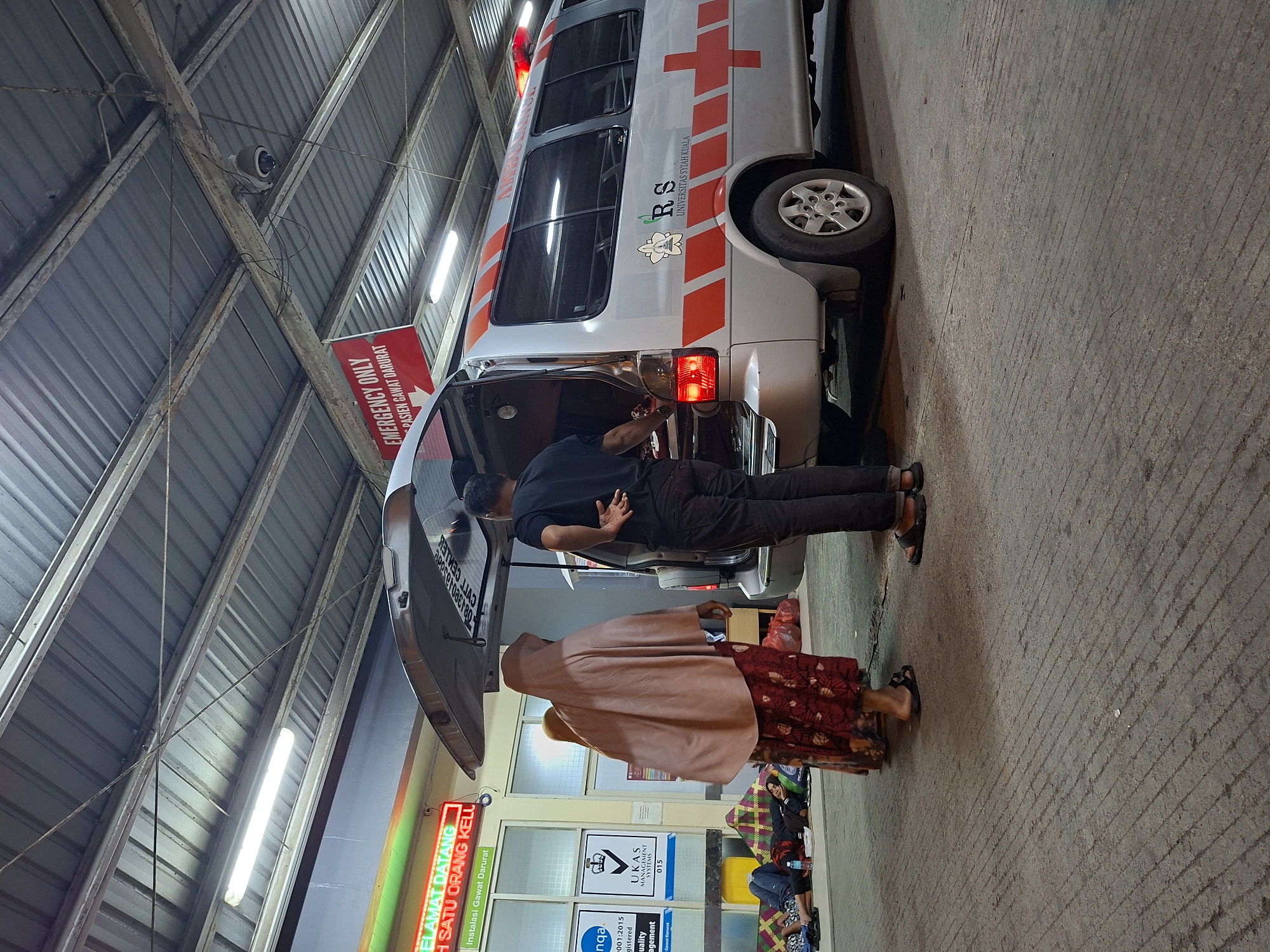 The image depicts an ambulance stationed outside a building, likely a hospital or clinic, identified by the "Emergency Only" sign in Indonesian and the presence of medical staff. The location is identifiable as Banda Aceh, Indonesia, based on the "Universitas Syiah Kuala" text on the ambulance. A man in a black shirt and dark pants is interacting with the open rear of the ambulance. Nearby, a woman wearing a hijab and a floral-patterned skirt stands, possibly accompanying a patient. Other individuals are seated on the ground against the building, covered with blankets or mats. The lighting suggests it might be evening or night. The building's architecture features a corrugated metal roof and bright fluorescent lighting.