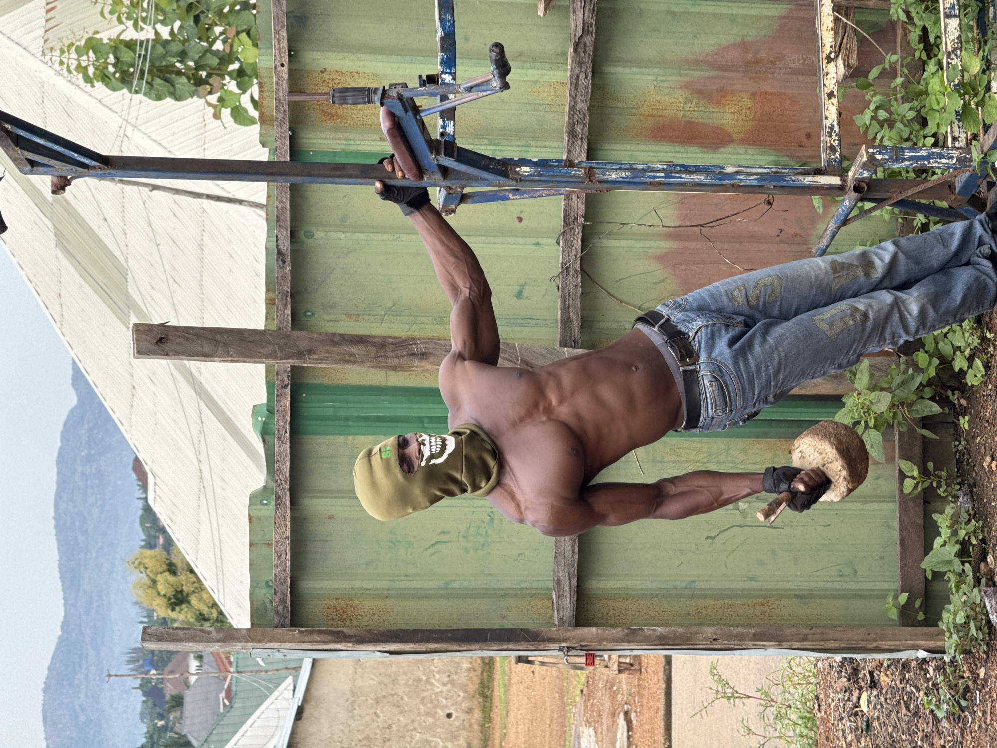 A shirtless man wearing a green balaclava with a white skull design and blue jeans stands outdoors in Akwanga, Nigeria, leaning against a blue metal fitness apparatus. He is holding a weight made of what appears to be a stone or concrete attached to a wooden handle in his gloved right hand. His left hand rests on the metal apparatus. The background features a corrugated green metal fence, a wooden post, and a white corrugated roof structure. Beyond that, buildings and hills are visible under a cloudy sky. The ground is covered with dirt, weeds, and some litter.