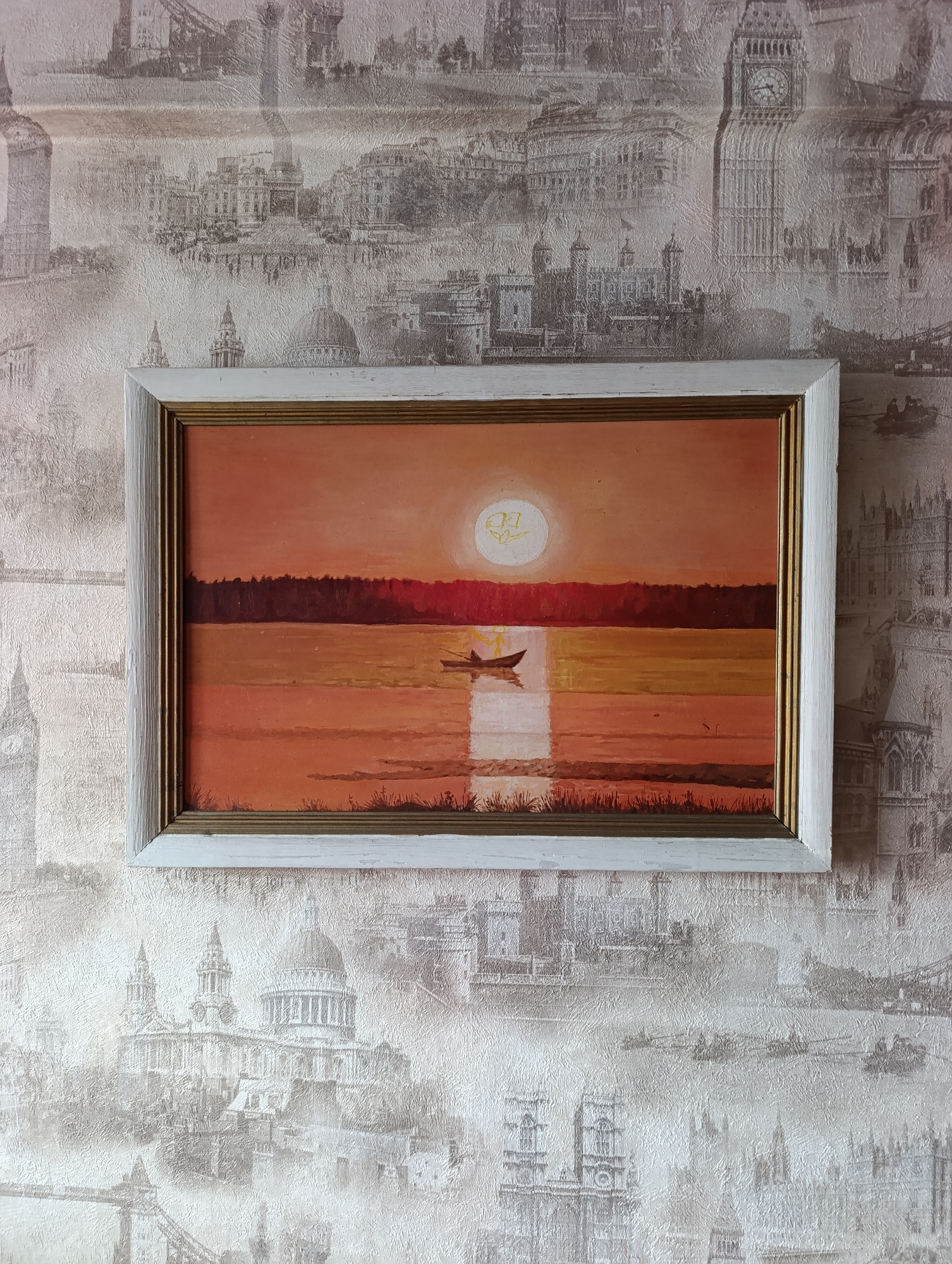 A framed oil painting hangs on a wall with London-themed wallpaper. The painting depicts a reddish-orange sunset over water with a dark line of trees on the horizon. A single boat is visible on the water in the center of the painting. In the sun, there is a symbol resembling a stylized butterfly. The painting has a white frame with a gold inner trim. The wallpaper behind the painting is a faded beige color with monochrome images of London landmarks, including Big Ben, the Tower Bridge, and St. Paul's Cathedral. No specific details indicate a location in Krasnoyarsk, Russia.
