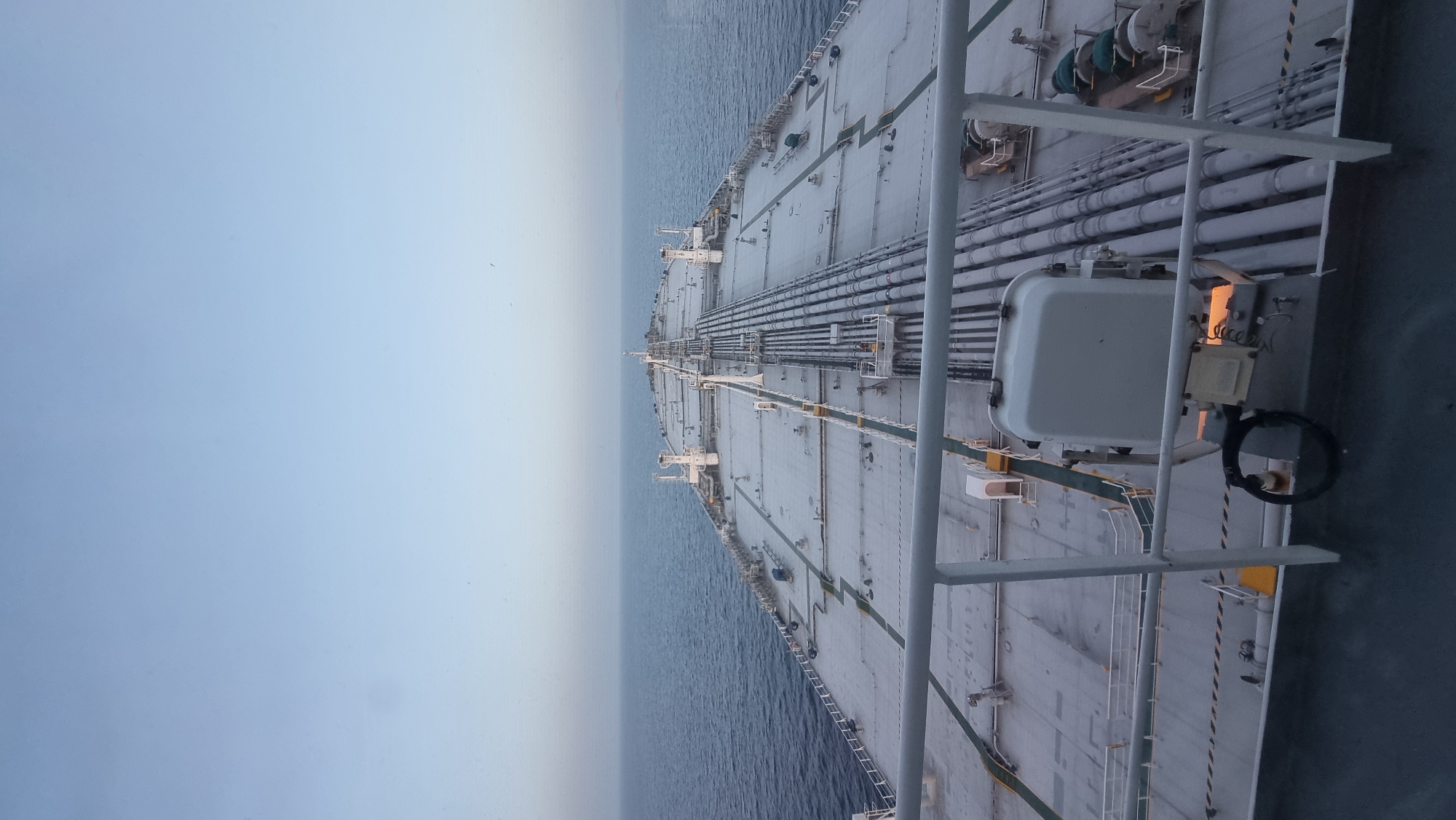 The image, taken from the deck of a large ship, captures a wide expanse of calm, blue ocean under a clear, pale blue sky. The perspective looks forward along the length of the ship, revealing a vast, flat deck covered in a light gray surface. Numerous pipes, running in parallel lines, dominate the central part of the deck. White railings run along the edges of the visible deck area in the foreground. In the distance, the ship tapers towards the horizon, where the sea and sky meet. There are no visible people or other objects on the deck, suggesting an unoccupied space. The environment is entirely maritime.