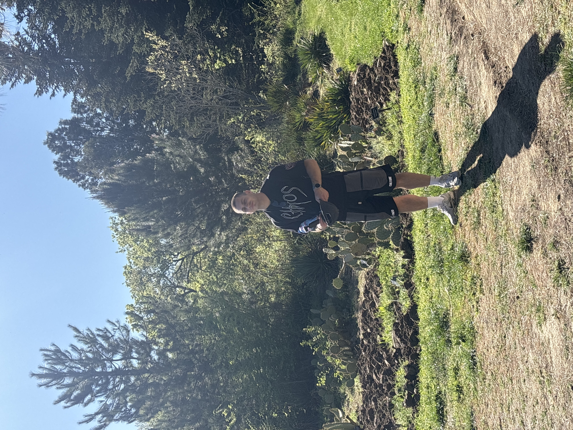 A full shot, portrait view of a light-skinned male standing outdoors on a bright day. He is in a grassy, natural area with trees and cacti in the background. He is wearing a black t-shirt with a white design, black shorts with lettering on the side, white socks, and grey athletic shoes. He holds a dark object in his left hand. His shadow is visible on the ground to his right. The location is potentially in a park or botanical garden, but the information is missing that it is in Varna, Bulgaria.