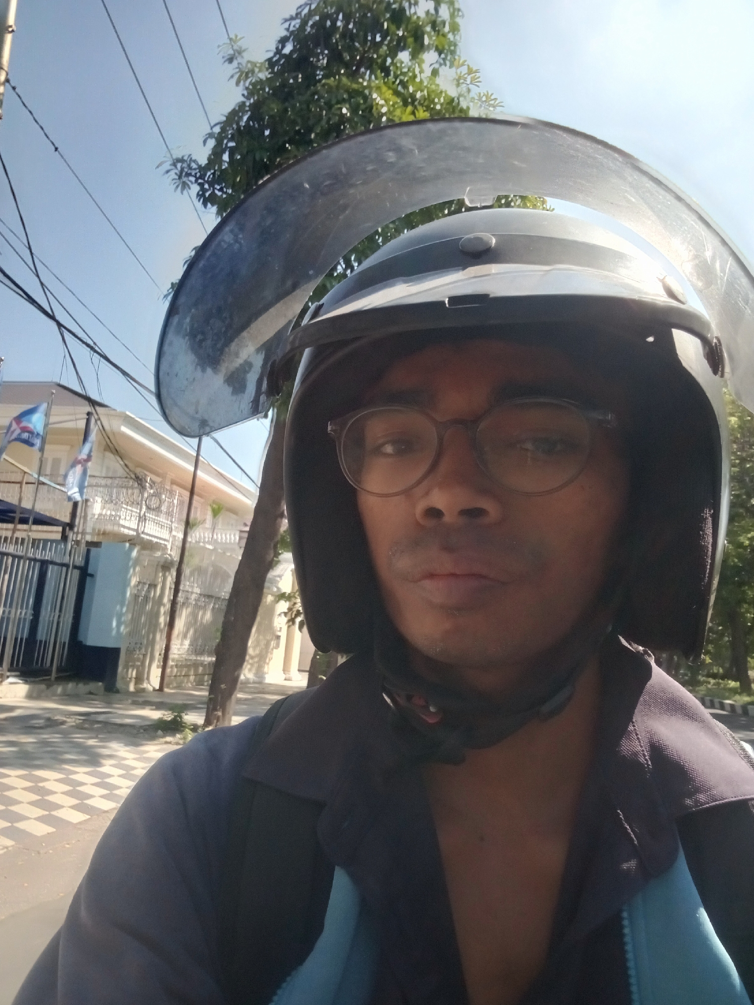 A person wearing a helmet with a clear visor, glasses, and a dark jacket is taking a selfie. The background shows a light-colored building with white railings, flags, and some trees. The pavement in front of the building has a checkered pattern of light and dark squares. Overhead, there are power lines against a clear blue sky. The overall scene suggests an outdoor urban environment, potentially in Surabaya, Indonesia, given the architectural style and vegetation. The person appears to be on a motorcycle or similar vehicle, indicated by the helmet and the perspective of the selfie.