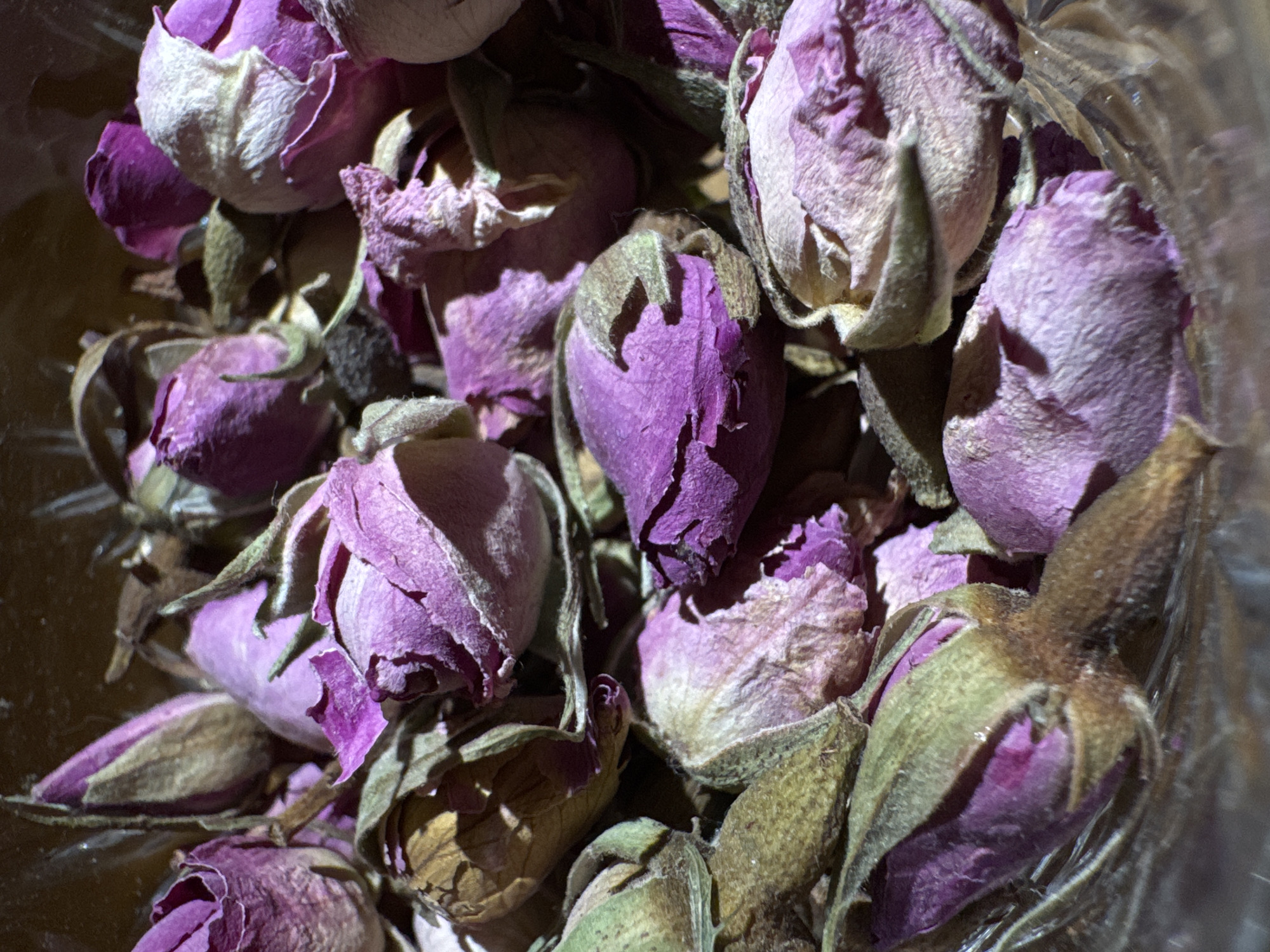 A close-up shot shows a collection of dried rosebuds, primarily in shades of purple and lavender, with some buds retaining hints of green and brown from their stems and sepals. The buds appear to be clustered together, potentially within a glass or container, judging by the slight distortion and highlights that could be reflections off a glass surface. The focus is sharp, highlighting the texture and detail of the dried petals. There is no indication of a specific location in Yerevan, Armenia, or any people, actions, or interactions.