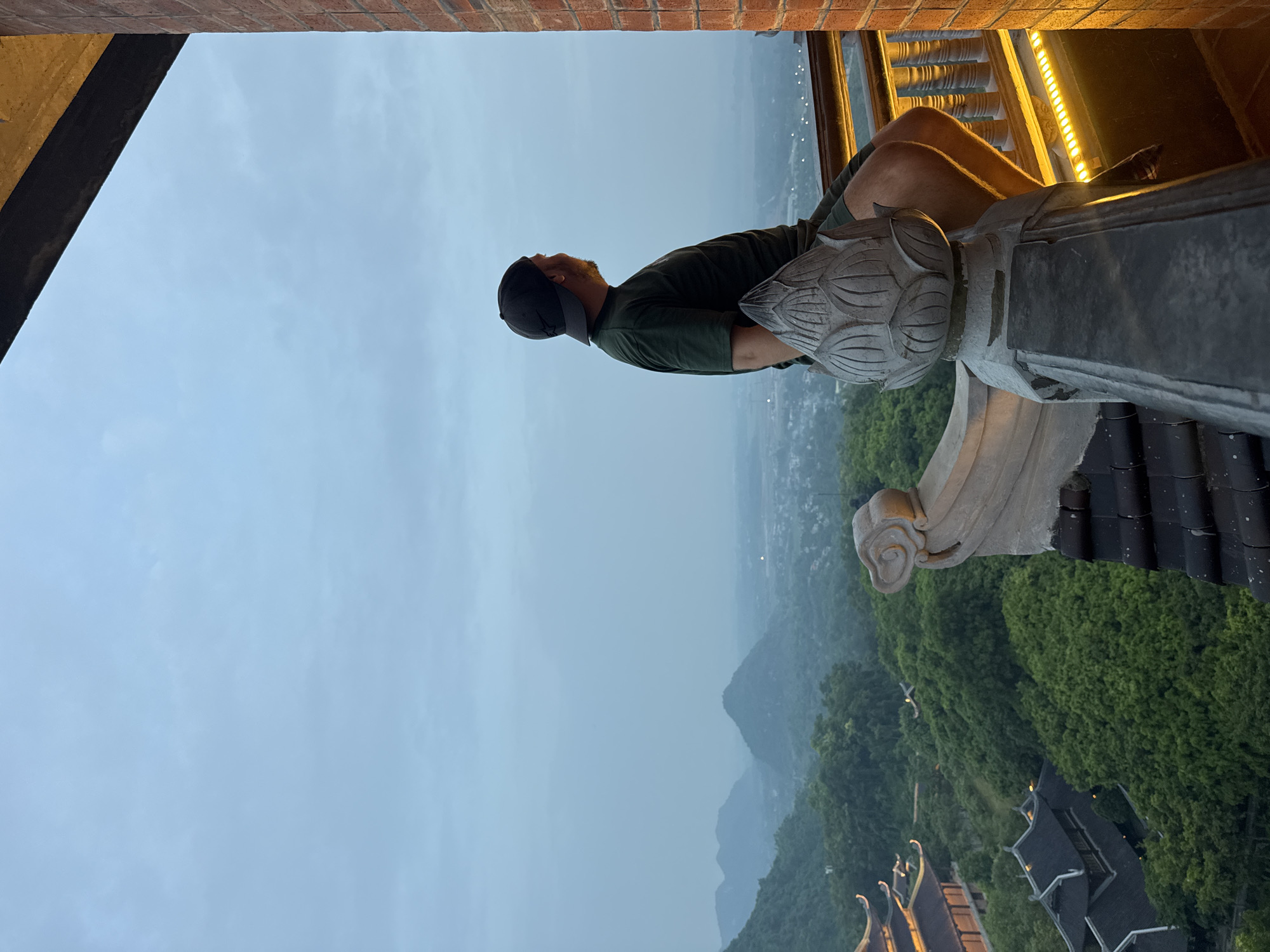 A man wearing a dark green t-shirt and a black baseball cap is sitting on a stone railing, looking out at a landscape. He is positioned on the right side of the frame, with his body turned away from the camera. The railing is ornate, featuring a carved lotus flower motif and a weathered, grey finish. Behind the man, a brick wall with a dark beam above it frames the scene. The background reveals a vast expanse of rolling green hills and mountains, with traditional Vietnamese architecture visible among the trees. The sky is overcast, with a soft, diffused light. The overall atmosphere is serene and contemplative. The location is identified as Tây Hoa Lư Ward, Vietnam.