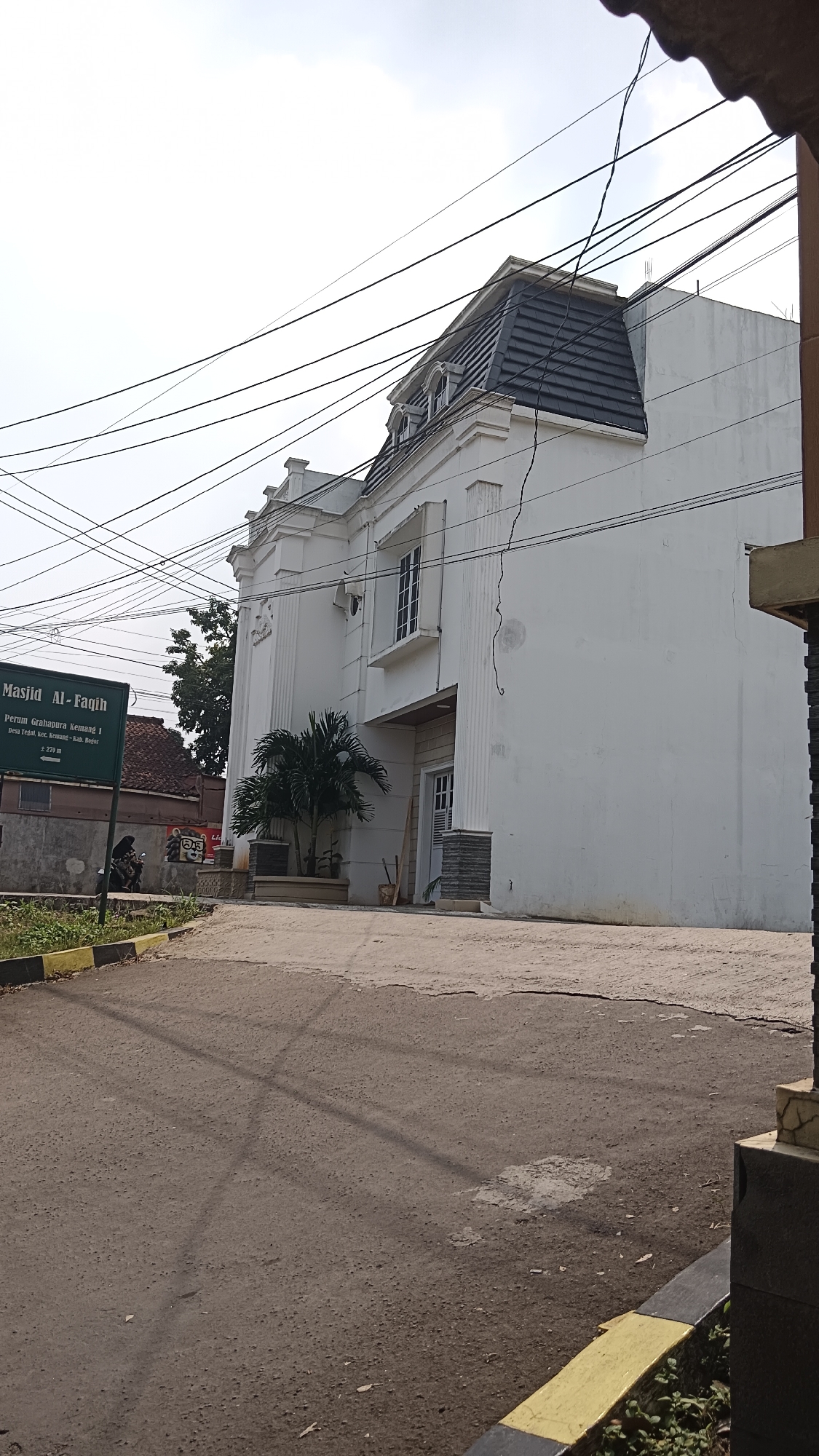 The image shows a street scene in Kemang, Indonesia, featuring a white building identified as "Masjid Al-Faqih" by a green sign. The building has a distinctive mansard roof with grey tiles and dormer windows. A small palm tree is planted in front of the building. The street in the foreground is paved and shows signs of wear. Electrical wires are prominently visible crisscrossing the sky above the building. To the left of the building, a portion of a residential house with a red roof is visible. A few people are present near the sign, one of whom appears to be riding a motorcycle. The environment suggests an urban or semi-urban setting with vegetation visible.