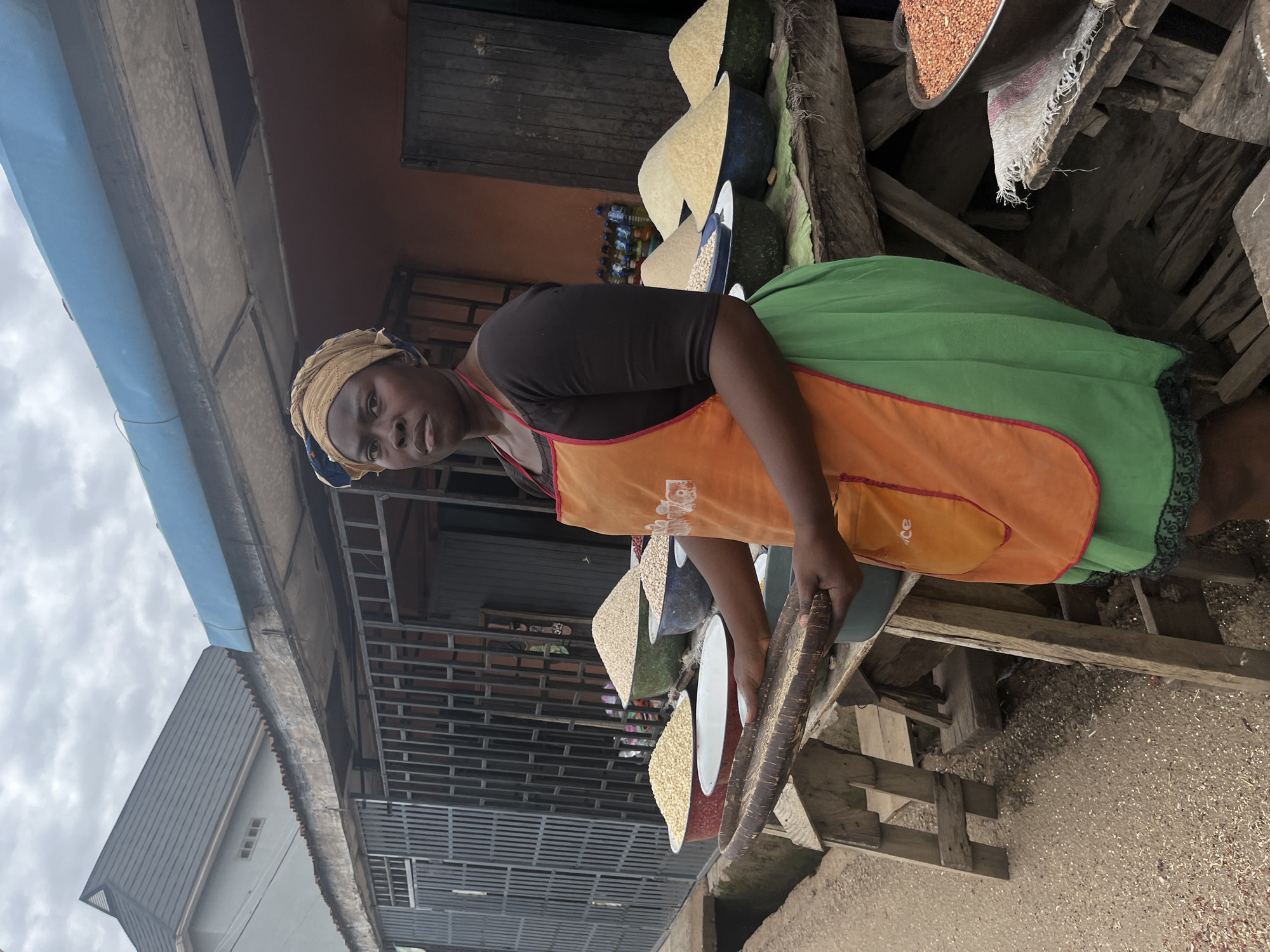 A woman stands in front of a roadside stall in Jos, Nigeria, where grains are displayed in bowls. She wears a headscarf, a dark shirt, an orange apron, and a green skirt. She holds two dark, wooden objects, possibly serving tools. The stall has multiple bowls containing different types of grains. The background includes buildings with metal structures and a cloudy sky. The ground is covered with scattered grain.