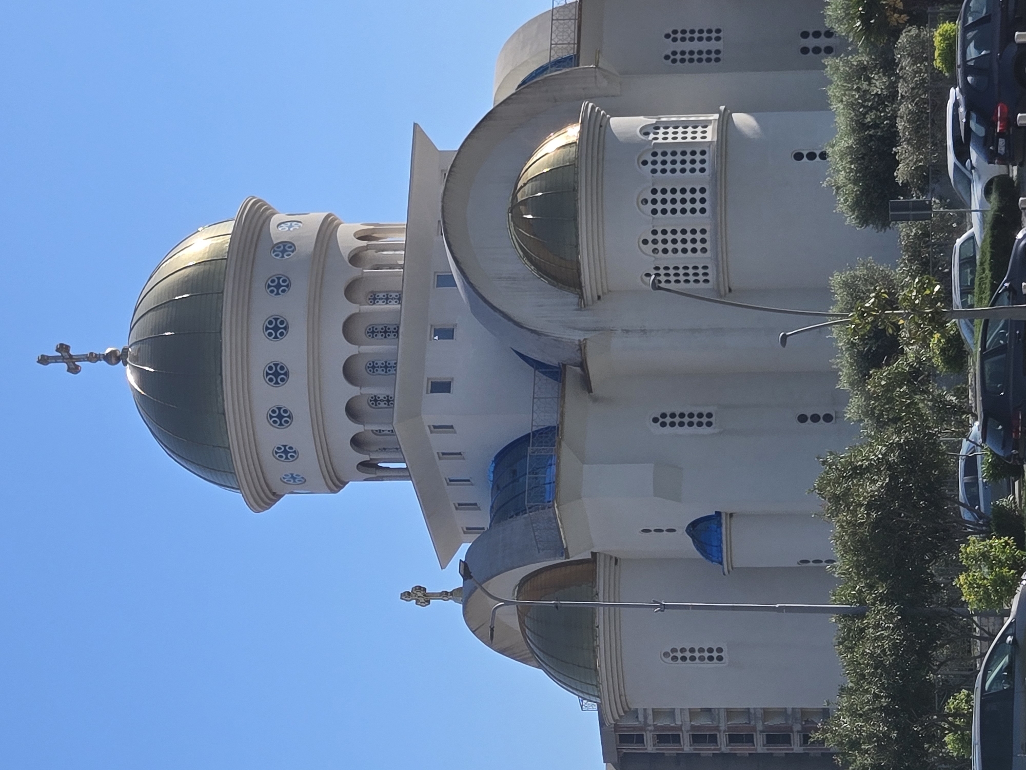 The image depicts a section of the St. Jovan Vladimir Orthodox Cathedral in Bar, Montenegro, taken from a low angle. The architecture features a large, golden-colored dome topped with a cross, and tiered levels with decorative windows. The building's exterior is primarily white. A clear blue sky is visible in the background. Olive trees are planted along a nearby street, with parked cars partially visible behind them. Streetlights are also present.