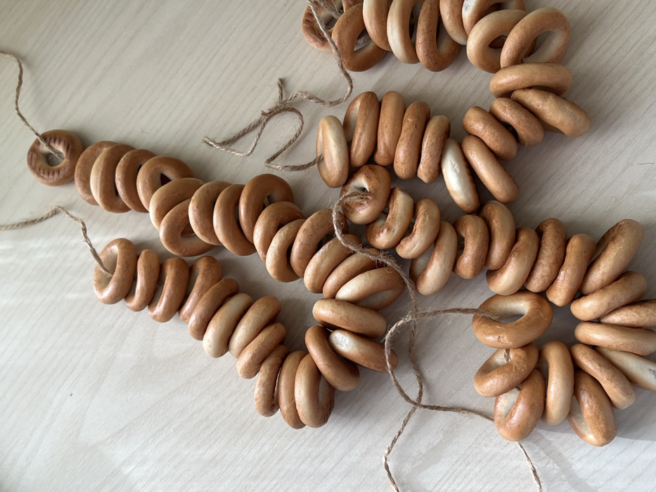 Four strings of bagels, also known as "bubliki," are arranged on a light surface. Each string is composed of multiple circular baked goods strung together using thin, natural twine. The bagels vary in color, ranging from light tan to a darker brown. The strings are positioned in a curved pattern, creating a visually appealing arrangement. The overall impression is that of a traditional, rustic food item.