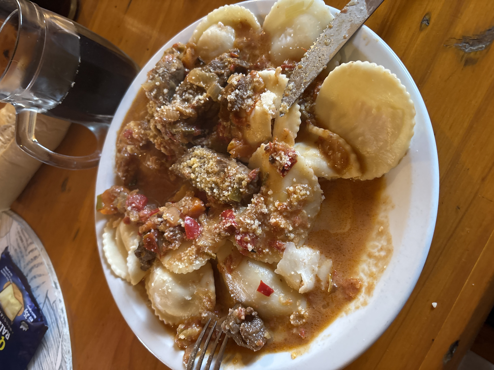The image shows a plate of ravioli with a meat-based sauce, garnished with what appears to be grated cheese. A fork and knife are present. The dish is on a white plate placed on a wooden table. A glass pitcher with a dark liquid and a package of food are visible in the periphery. Based on the metadata, the image was taken in Villa General Belgrano, Argentina.
