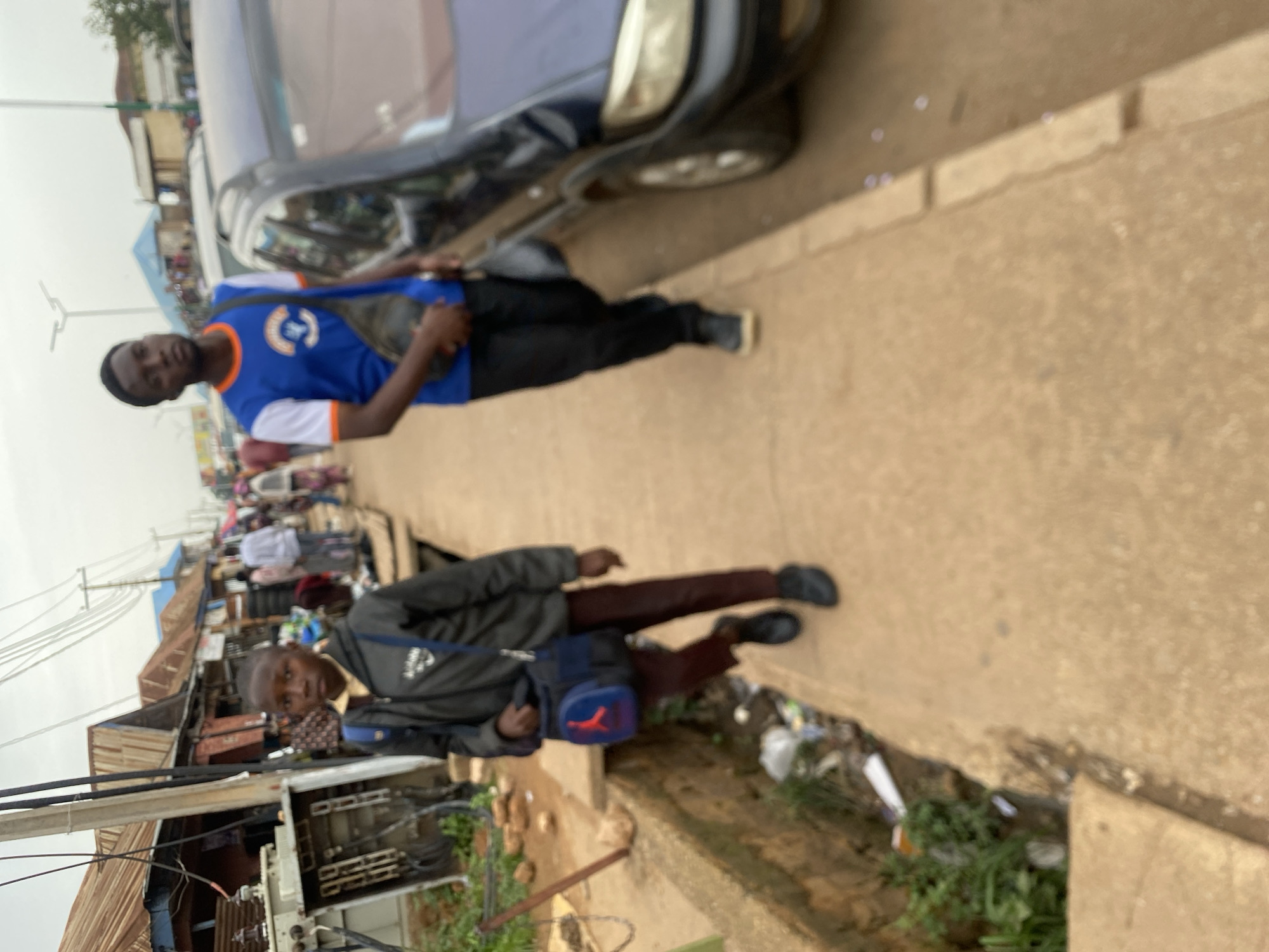 In Bukuru, Nigeria, a young boy wearing a dark jacket and maroon pants walks down a paved sidewalk. He carries a dark bag with a blue and red logo. Behind him, a young man in a blue and white shirt with orange trim and black pants walks, also carrying a bag. They are in a busy area with market stalls and vehicles on the side of the road. The environment appears to be a typical urban street scene, with overhead wires and some litter on the side of the sidewalk. The sky is overcast.
