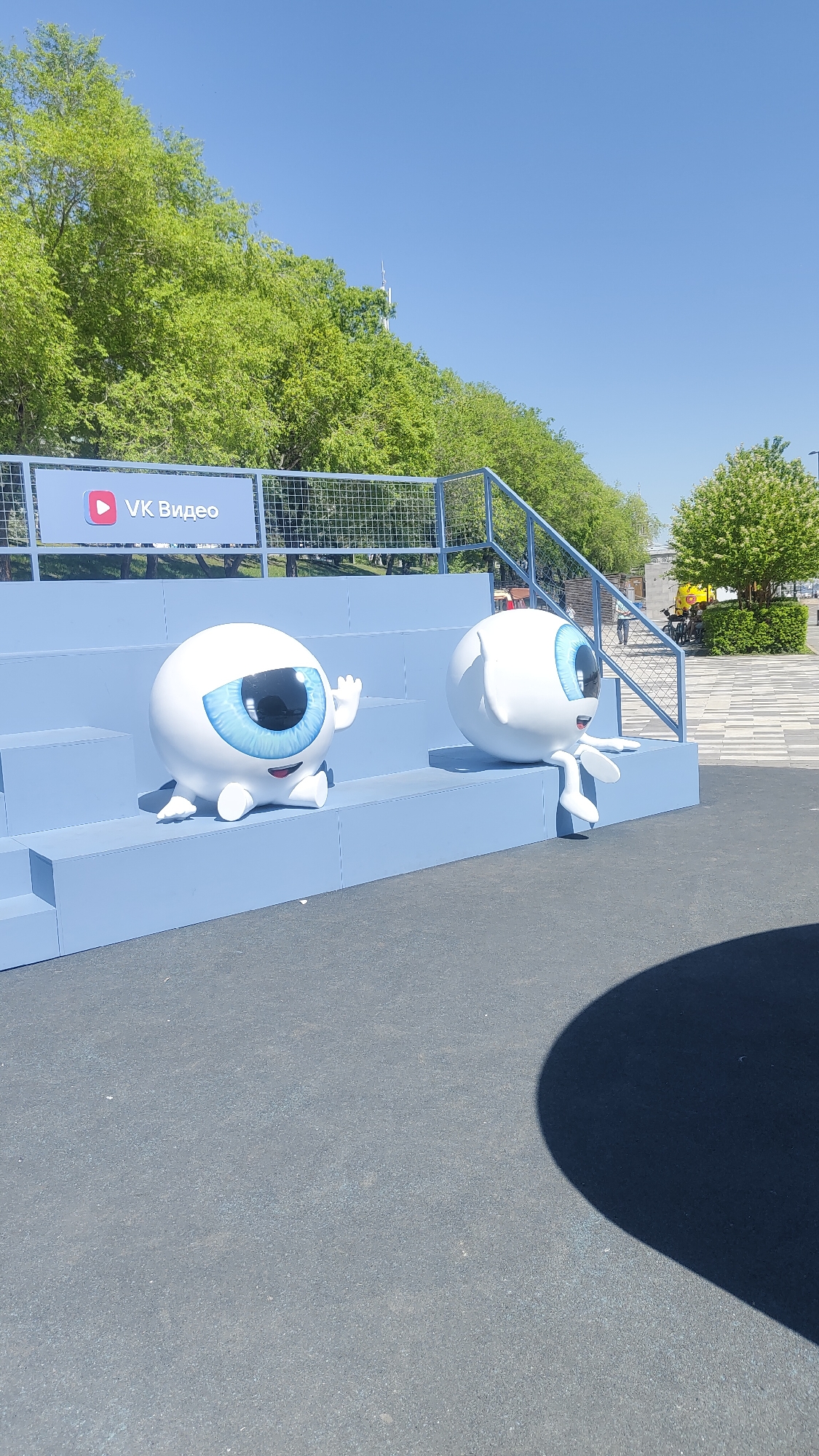 This outdoor image, likely taken in Krasnoyarsk, Russia, on a bright, sunny day, features two large, white, spherical mascots resembling eyeballs, displayed on a light blue tiered seating structure. The mascot on the left is seated, facing forward, with a prominent blue iris and pupil, a smiling mouth, and its right hand raised in a wave. The second mascot, to the right, is also spherical with a blue eye and a smiling mouth, seated and facing left with its legs dangling from a lower tier of the seating.
Behind the blue tiers, a metal fence is visible, bearing a sign with the red play button logo and the text "VK Видео" (VK Video), indicating a connection to the Russian social media and video platform. Lush green trees form a dense backdrop, suggesting a park or natural area. The sky above is clear and blue. The foreground consists of dark grey paved ground, with a large, dark, curved shadow extending from the lower right corner, indicating strong sunlight. In the distant background, faint figures of people and a yellow structure can be discerned. The overall scene appears to be a public installation or event setup, conveying a cheerful and inviting atmosphere through the mascots' expressions and bright weather.