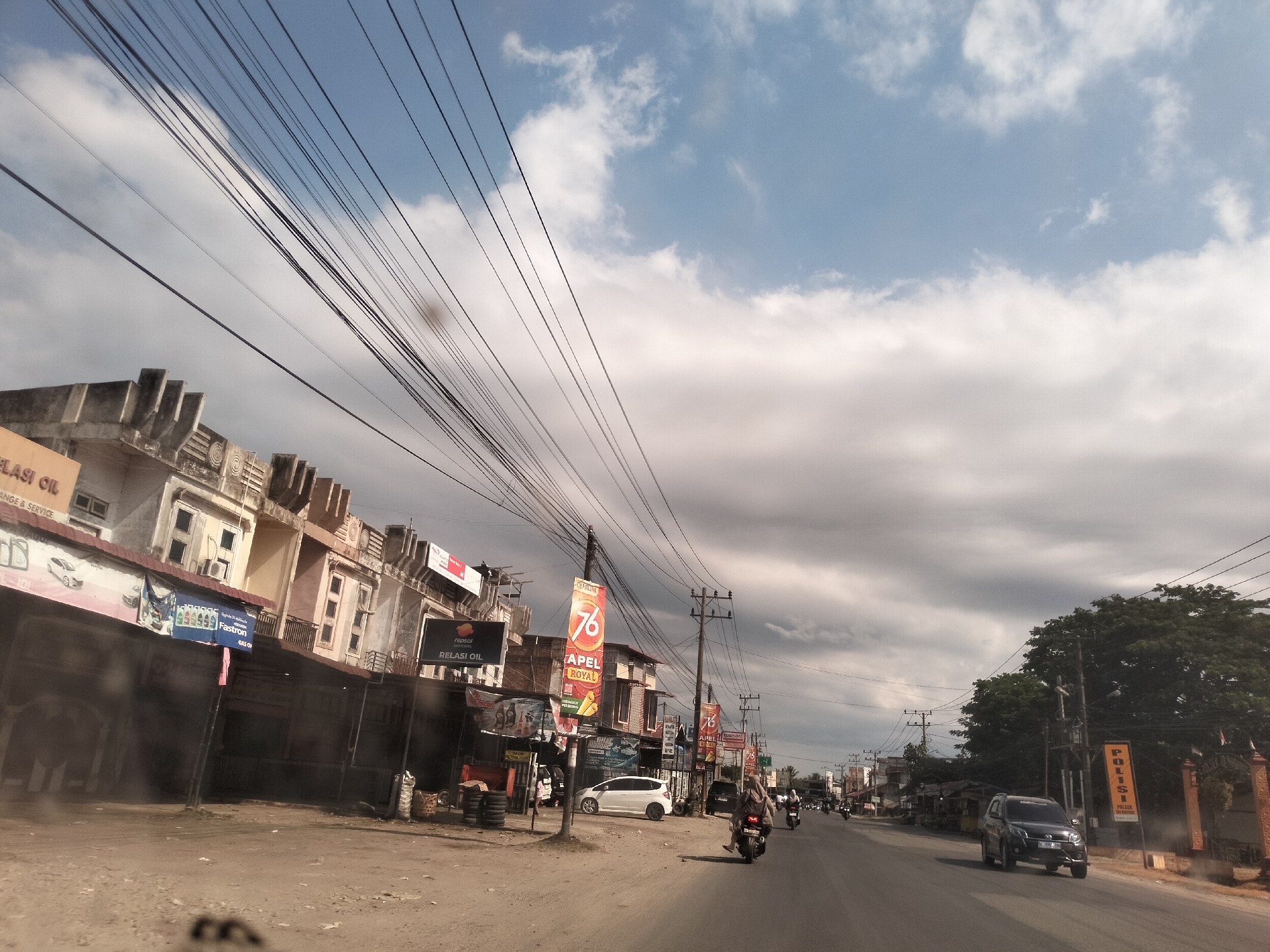 The image shows a street in Teupinbatee, Indonesia. The street has a road and a sidewalk. On the sidewalk, there are buildings, signs, and trees. The signs advertise gasoline, including a "76" sign. There are multiple vehicles on the road, including cars and motorcycles. Overhead, there are numerous electrical wires. The sky is partly cloudy.