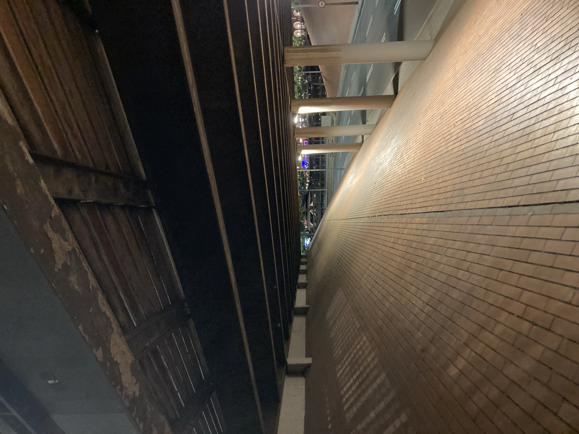 The image shows an elevated walkway or pedestrian bridge at night in Barcelona, Spain. The walkway is paved with light-colored brick or stone and slopes downwards to the right. It is covered by a dark ceiling supported by numerous parallel wooden beams, some of which show signs of wear and tear.
On the right side of the walkway, there is a road with a lane marking and streetlights visible in the distance. Several concrete pillars support the structure above the road. The area is illuminated by artificial lights, casting shadows on the walkway. There are no people visible in the image. The overall scene suggests an urban infrastructure at night, possibly a bridge over a road or a pedestrian path in a city.