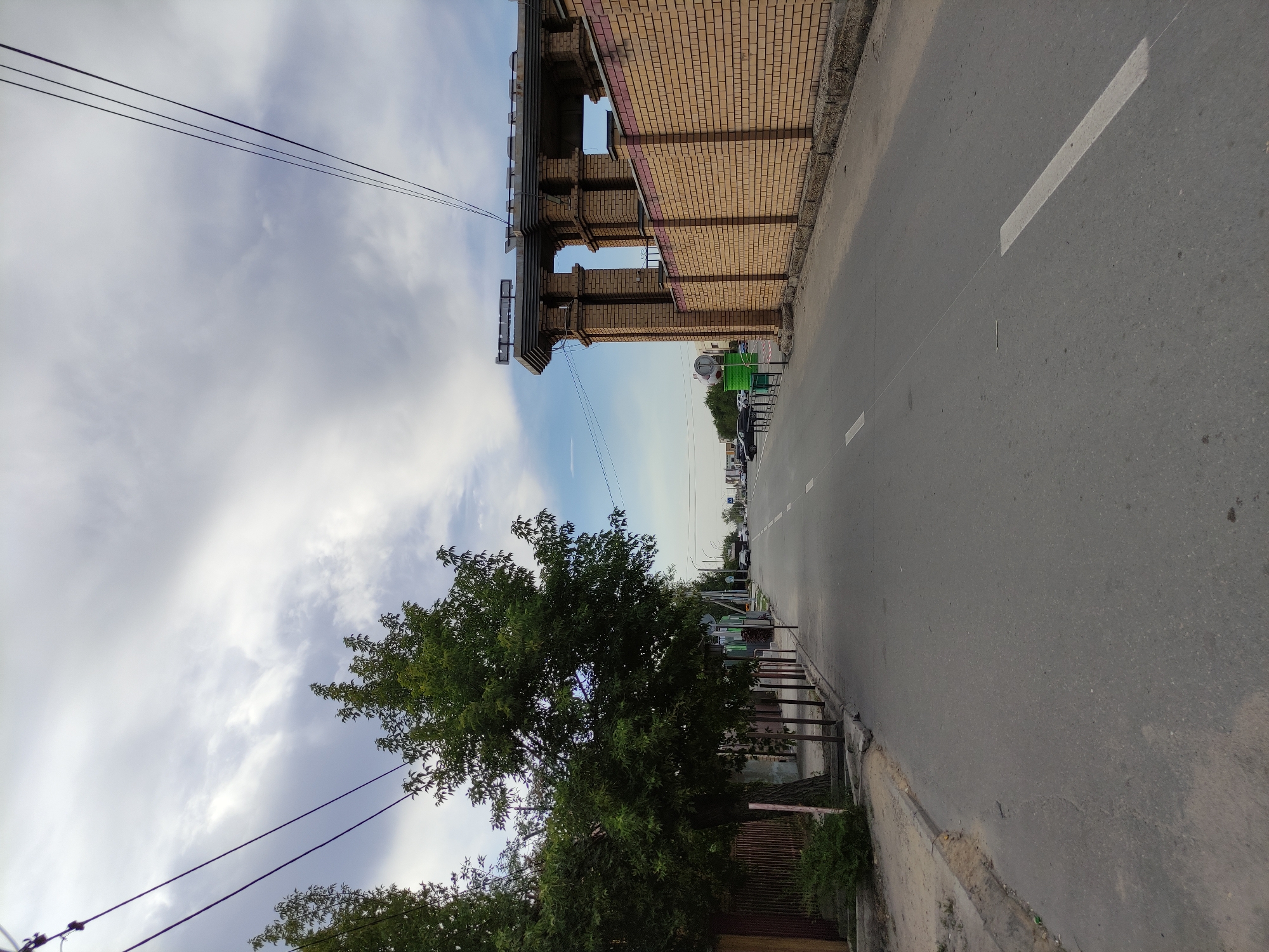 The image depicts a street scene in Semey, Kazakhstan, with a focus on a paved road running through an urban environment. On the left side of the road, a sidewalk is visible, bordered by a fence and trees. The trees are dense with green foliage, and a few thin posts are planted along the sidewalk. On the right side of the road, a brick wall with a decorative cornice runs along the property line. The wall is constructed of light brown bricks. Beyond the wall, a building structure with columns and a decorative roofline is visible.
The road itself is asphalt, with white lane markings. Cars are visible in the distance, driving along the road. The sky above is overcast with scattered clouds. Power lines are strung across the top of the image, indicating the presence of electrical infrastructure. The overall impression is a typical street scene in a city, with elements of architecture, nature, and urban infrastructure present.