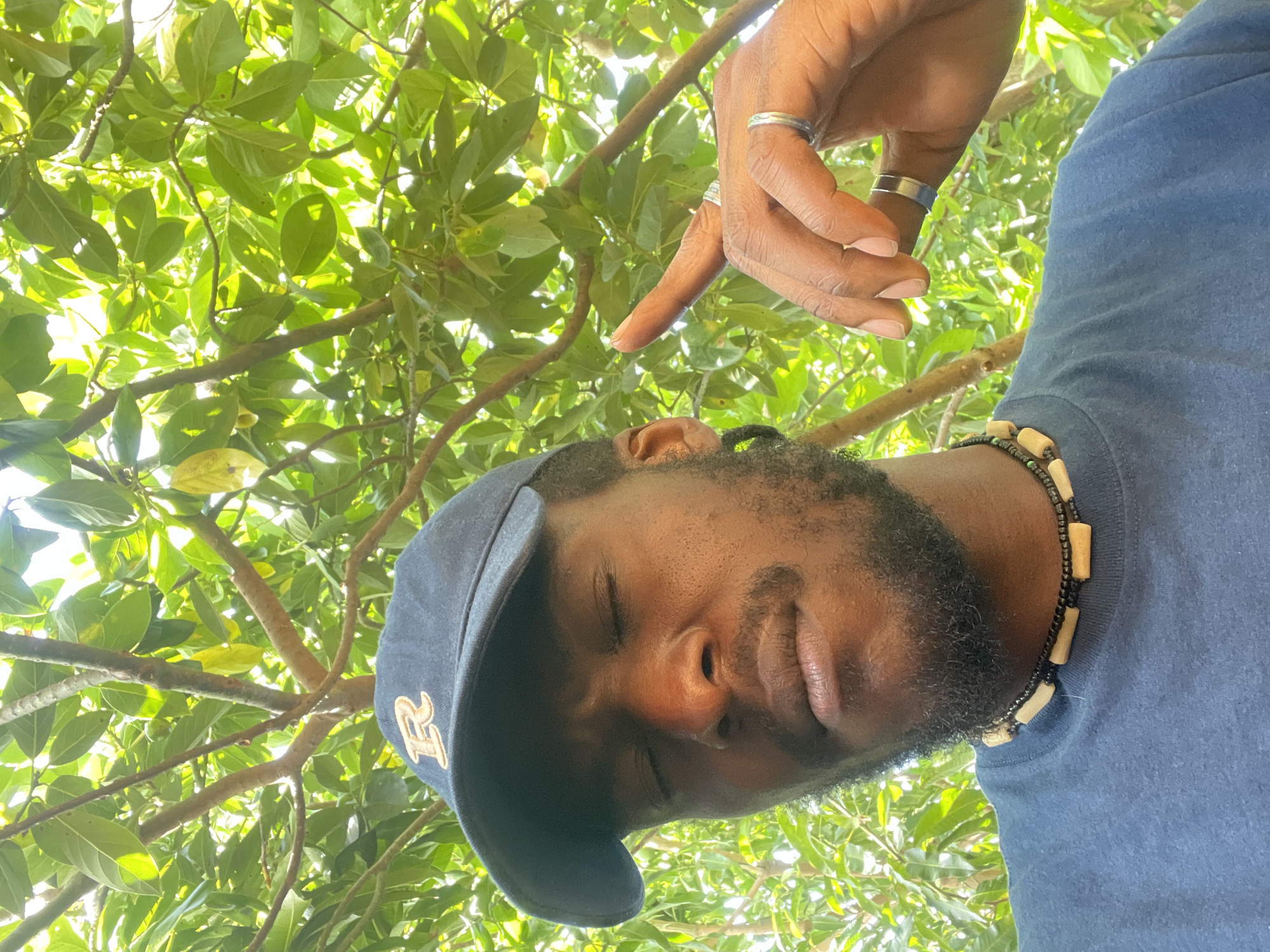 A low-angle shot shows a dark-skinned man wearing a navy blue baseball cap with a yellow "R" emblem. He has a beard and is wearing a necklace with beige cylindrical beads and smaller black beads. His eyes are closed, and his face appears serene. Above him, a hand with multiple rings on the fingers points down. The background consists of branches and leaves of a tree, illuminated by sunlight. There is no way to determine the location is the city of Jos, Nigeria from the contents of the image.