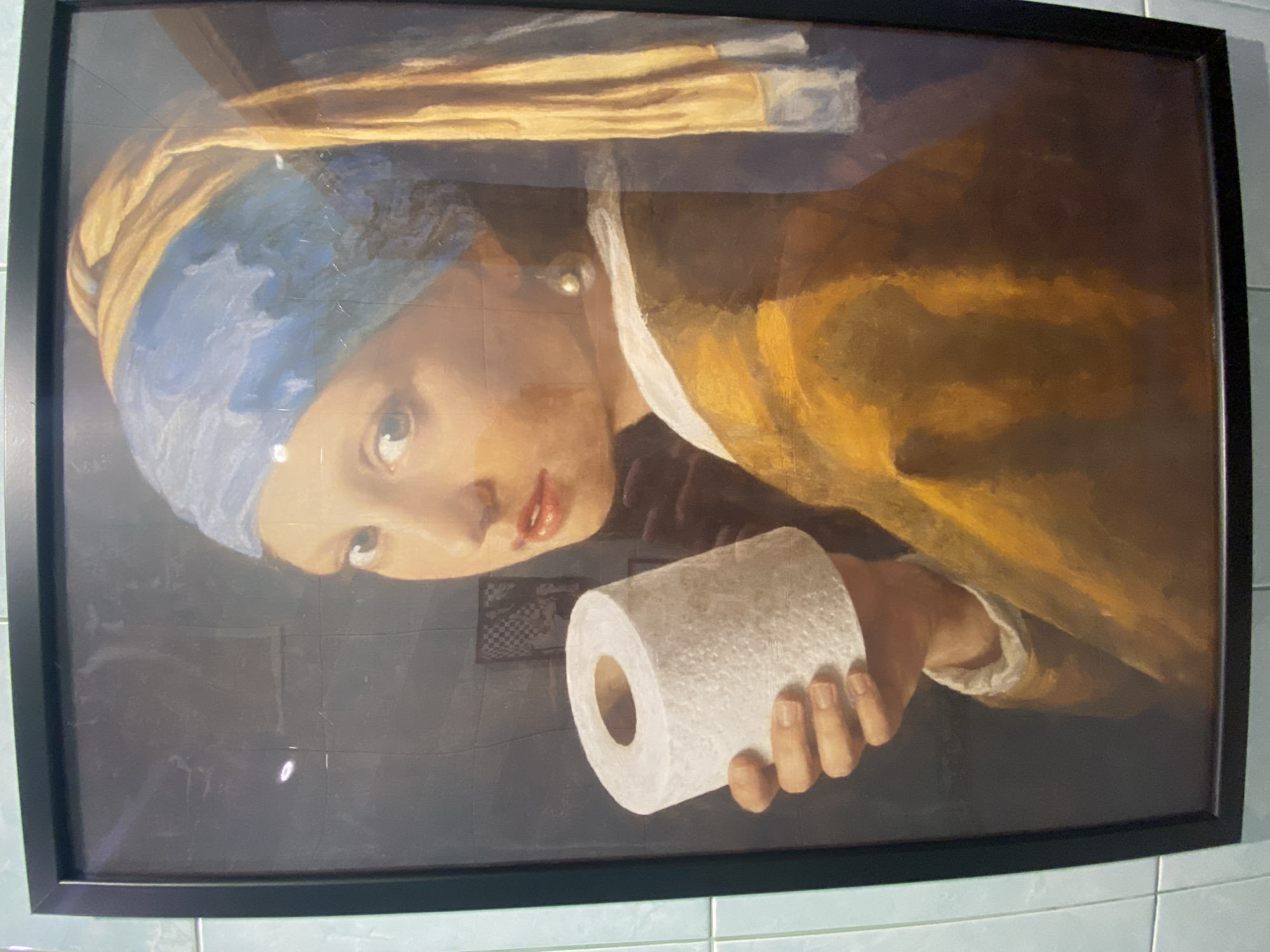 A framed image of "Girl with a Pearl Earring" by Johannes Vermeer, digitally altered to include the subject holding a roll of toilet paper, is mounted on a white tile wall. The black frame is visible, and the image is slightly rotated. The location is Krasnoyarsk, Russia.