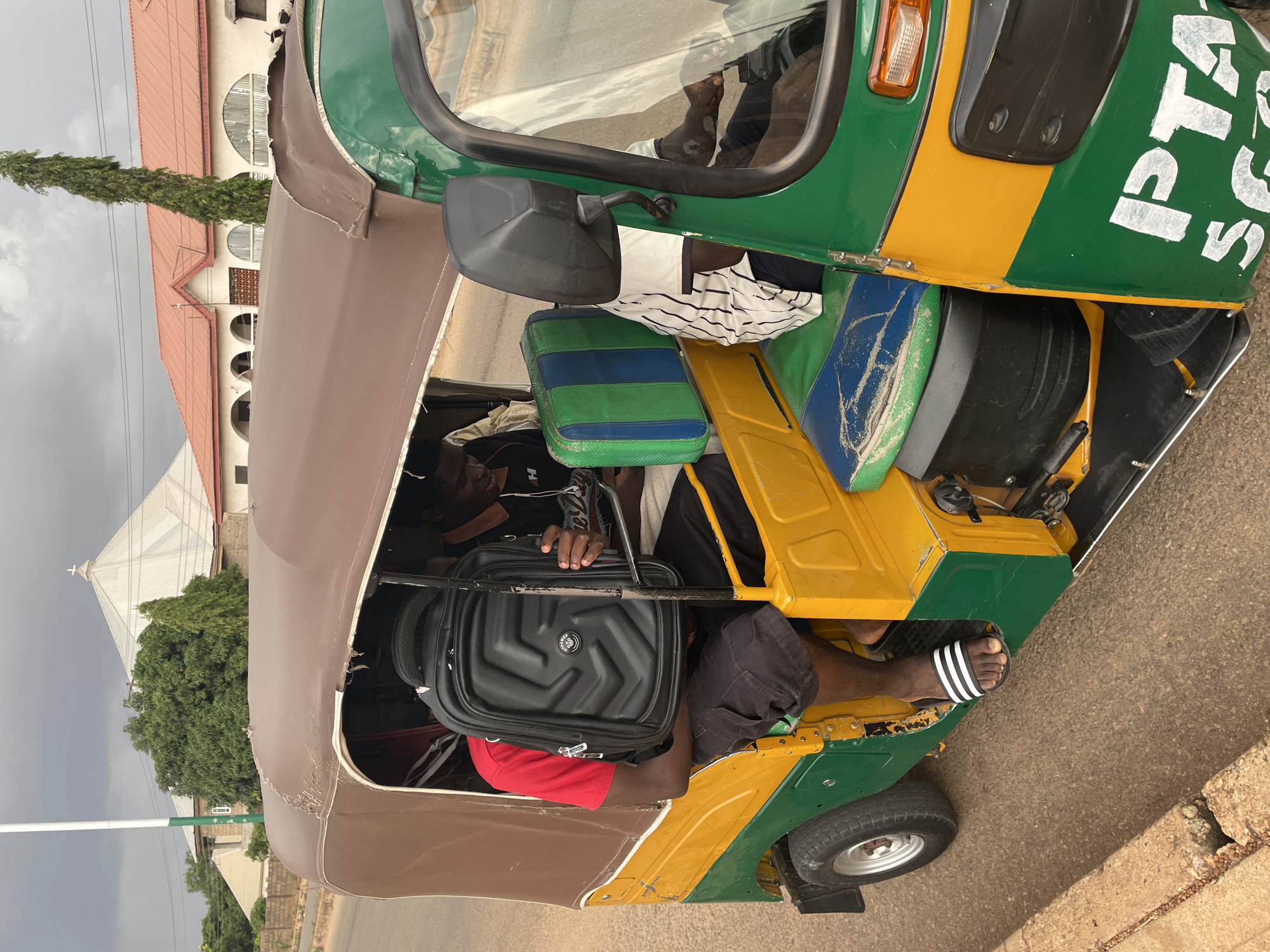 The image depicts the interior of a commercial tricycle in Jos, Nigeria, identifiable by the green and yellow paint scheme typical of "keke napep" transport vehicles. The tricycle is partially filled with passengers and luggage, including a large black suitcase held by one of the passengers. One passenger is wearing Adidas-branded sandals. The background shows a building with a red roof and arched windows, as well as a church with a cross, suggesting a residential or commercial area in the city. The weather appears overcast. The image is rotated counter-clockwise.