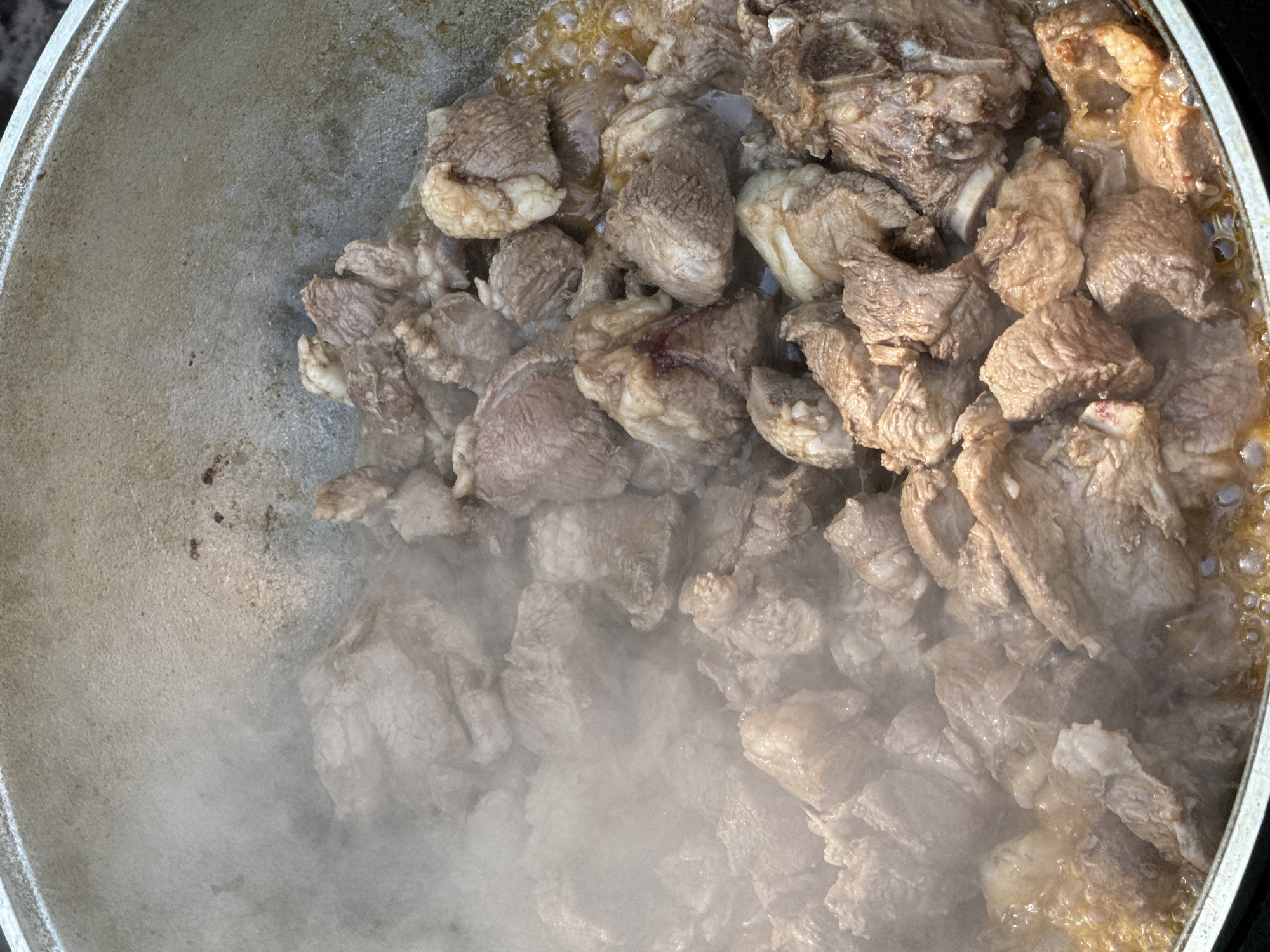 A close-up, overhead shot shows chunks of raw meat cooking in a large, metallic pot. The meat is various shades of brown and pink, with some pieces showing fat marbling. Steam rises from the pot, obscuring some of the meat. The pot appears to be made of cast iron or aluminum and has a worn, textured surface. The scene takes place outdoors, with a glimpse of a dark surface at the bottom edge of the frame, possibly a table or ground. The image depicts a cooking scene, possibly a traditional dish being prepared in Sary-Oy, Kyrgyzstan.