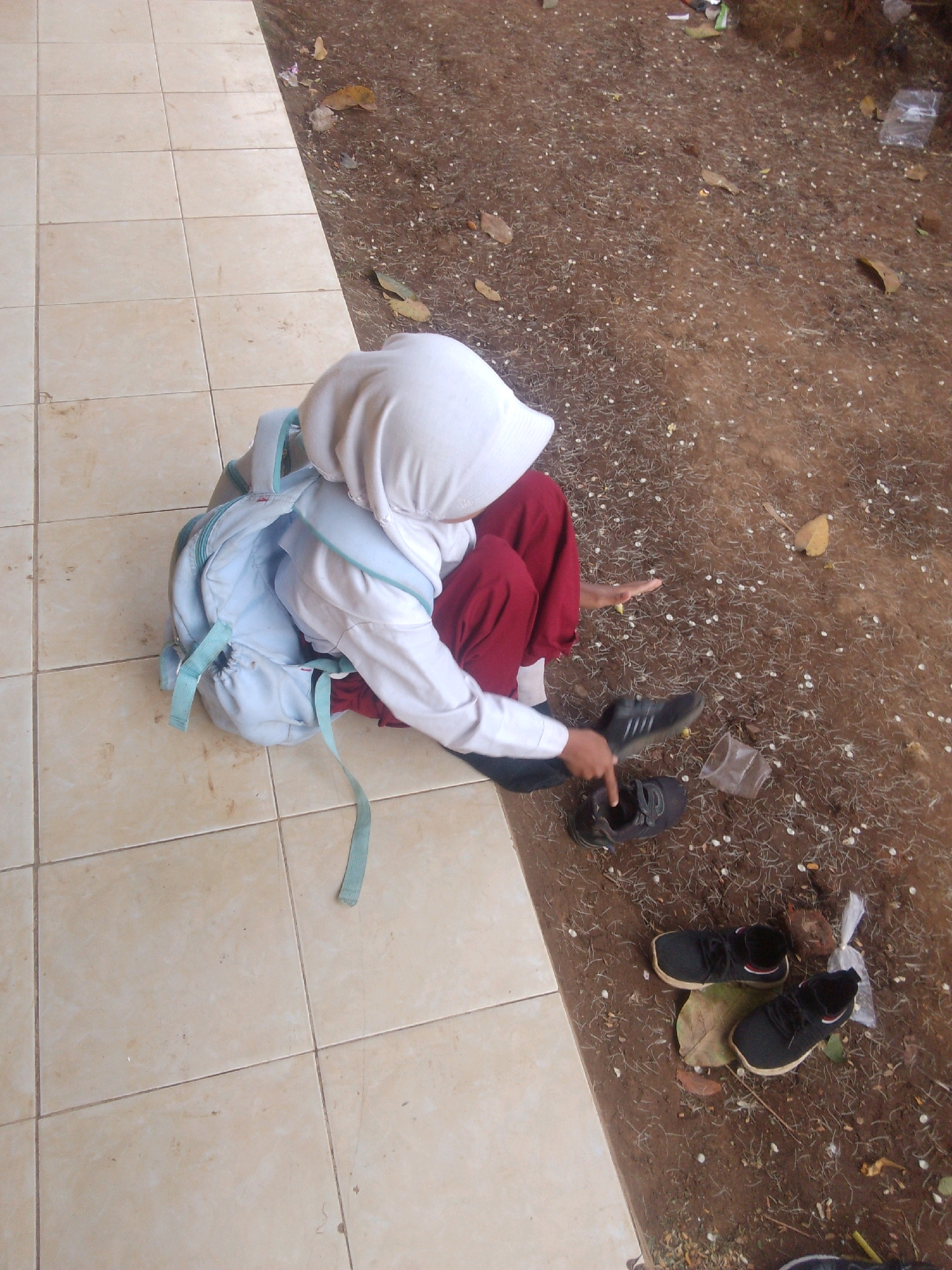 A young individual wearing a white headscarf, white long-sleeved shirt, and red trousers is seated on a tiled surface next to a dirt area. A light blue backpack is on their back. One shoe, a black sneaker, is on the individual's right foot, while another black sneaker is lying on the ground to their left, with the individual pointing towards it. A second pair of black sneakers sits on the dirt area to the right, next to a dried leaf and a plastic bag. The environment appears to be outdoors, possibly a schoolyard, given the tiled walkway bordering a patch of earth with scattered debris. The location is identified as Betek, Indonesia.