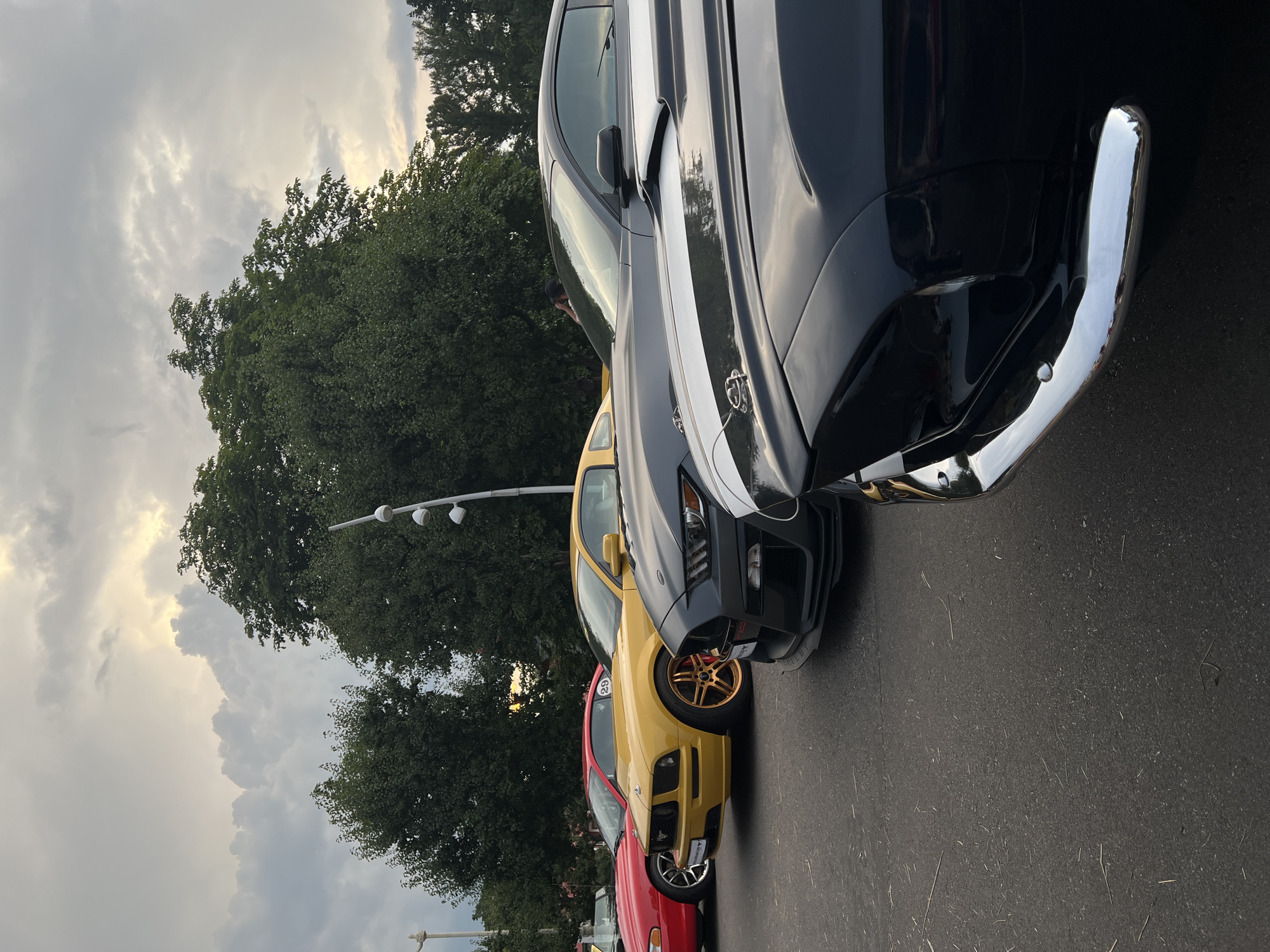 A line of Ford Mustang cars is parked on asphalt. The cars are, from front to back, black with white stripes, dark gray, yellow with gold wheels, and red. Trees and a street lamp are visible behind the cars. The sky is cloudy and light is visible through the clouds. The location is Moscow, Russia.