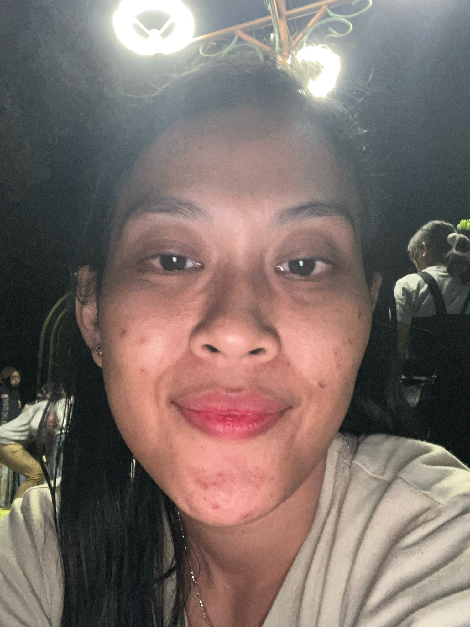 A close-up, selfie-style, horizontal, medium shot shows a woman's face filling most of the frame. She is outdoors at night, indicated by the darkness of the background and a bright overhead light source that is partially visible at the top of the image. The woman has dark hair that is pulled back, and she is looking directly at the camera with a slight smile. Her skin has some blemishes on her chin and cheeks. She is wearing a light-colored t-shirt. In the background, out of focus, are other people and structures that suggest a public gathering or outdoor event. Based on the image and the provided location, this scene is likely taking place in a public space within Kecamatan Pati, Indonesia.