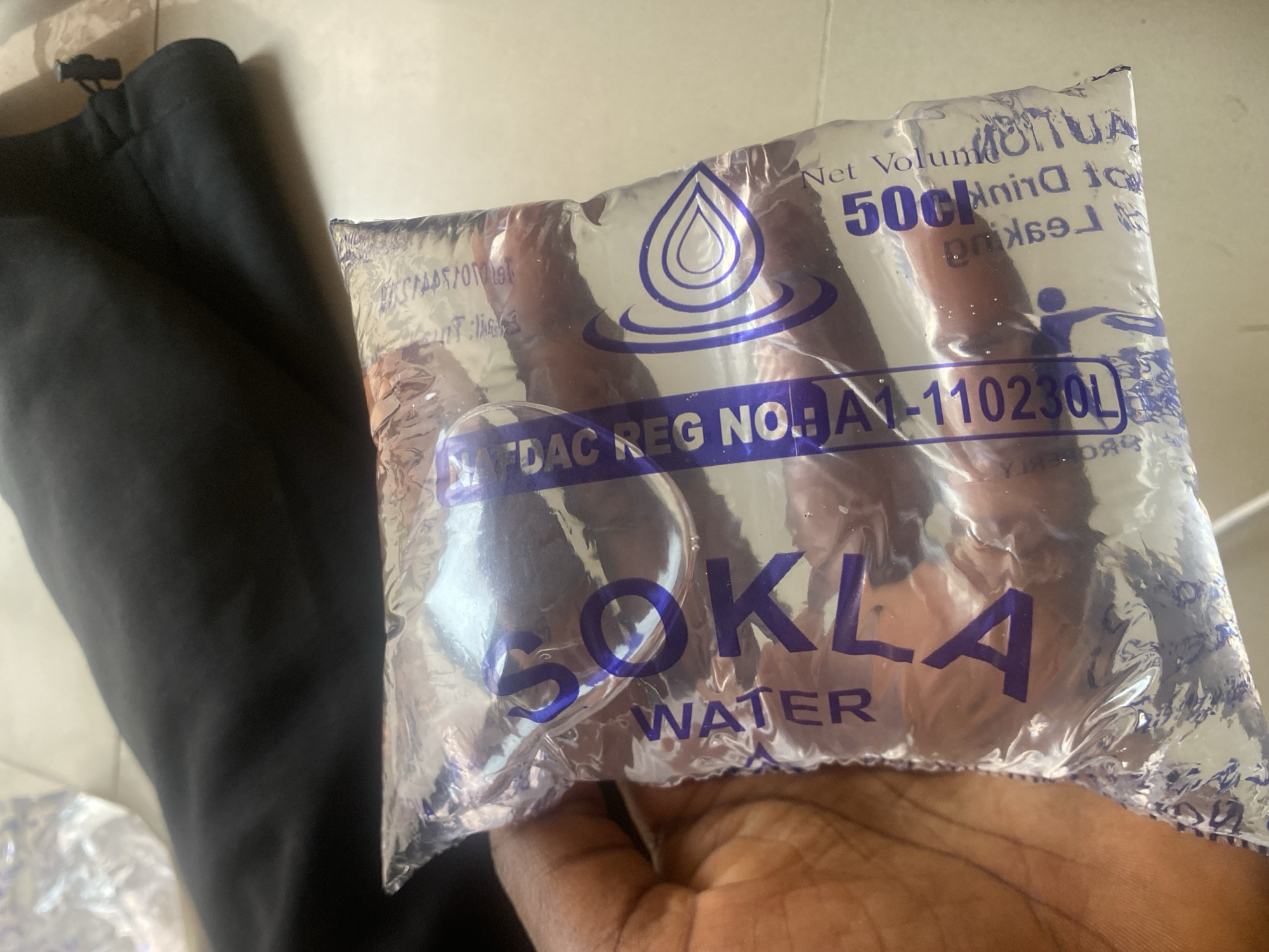 A person's hand is holding a clear plastic sachet of water. The sachet is filled with water and has purple text and graphics printed on it. A black piece of fabric, possibly clothing, is visible to the left of the sachet. In the background, the surface appears to be a light-colored tiled floor. The image has a bright and natural light source, suggesting it was taken during the daytime, possibly outdoors or in a well-lit indoor space. The overall scene is simple and focuses on the water sachet.
Visible text on the sachet includes:
- "SOKLA"
- "WATER"
- "Net Volume 50cl"
- "DO NOT DRINK TO LEAKAGE"
- "NAFDAC REG. NO: NA1-110230L"