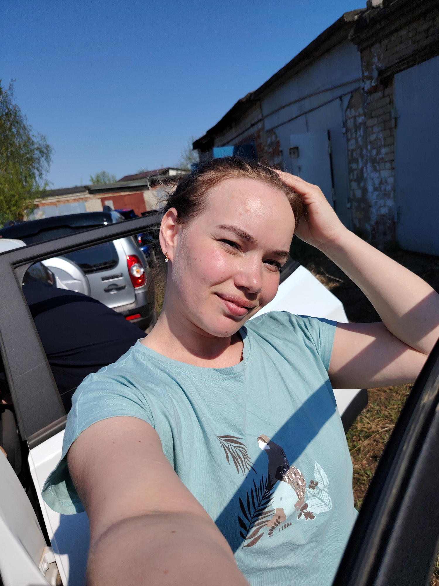 A light-skinned woman with auburn hair pulled back in a messy bun is taking a selfie. She wears a light blue t-shirt featuring a stylized graphic. She's in the open doorway of a white car. Behind the car are other parked cars, including a silver Lada. In the background is a one-story brick and concrete building with metal doors, typical of industrial or garage areas. The sky is blue and clear, suggesting a sunny day. Based on the surrounding details, the location is likely an industrial or garage area, perhaps in the city of Chaikovsky, Russia.