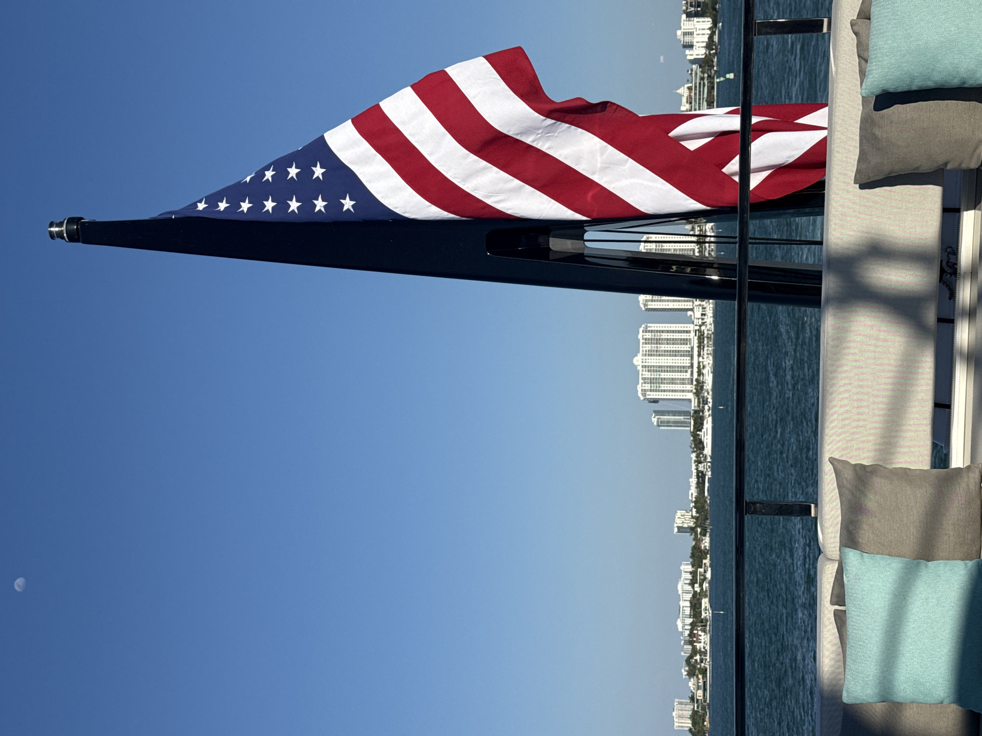 A sideways, wide shot features an American flag waving from the back of a boat with the Miami Beach skyline in the distance. The flag is attached to a black pole and has its blue star field on the left side of the image, with the red and white stripes flowing to the right. The water is calm and blue, with tall buildings lining the coast. A section of outdoor seating with gray and turquoise pillows is visible on the right side of the image. A small moon can be seen in the sky on the bottom left.
