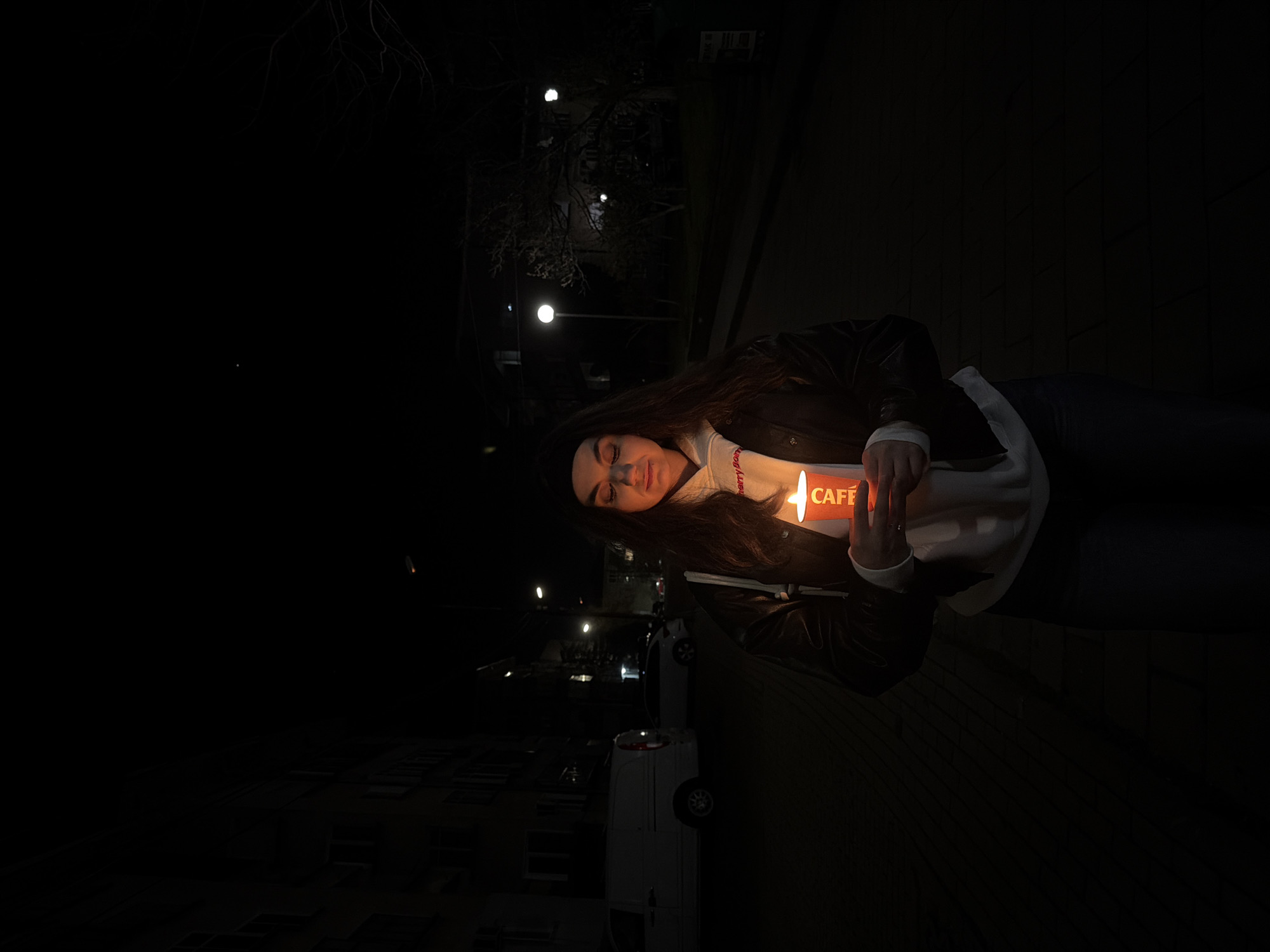 A woman is holding a lit "CAFE" sign in the dark. She is wearing a black jacket, white hoodie and a black beanie. The background includes a wall and a street with street lights and a white vehicle. There is no indication that the photo was taken in Karlovo, Bulgaria.