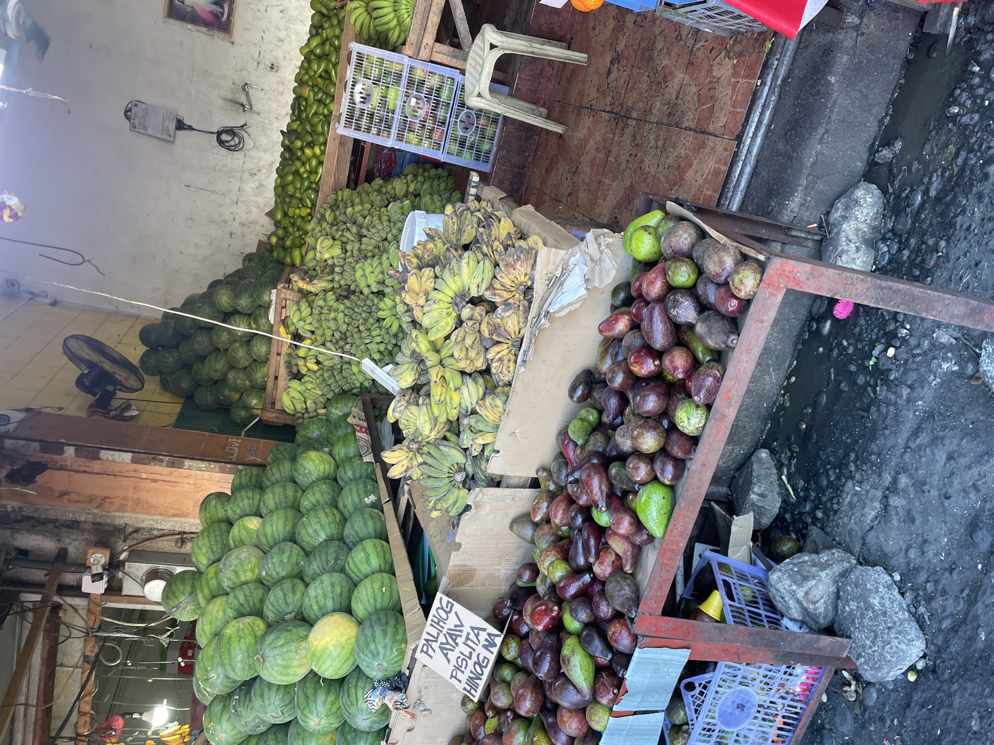 This image captures an outdoor fruit stall in Davao City, Philippines, primarily featuring large quantities of watermelons, bananas, and avocados. In the foreground, numerous avocados, varying in shades of green and purple-red, are displayed on a red-framed wooden stand. A handwritten cardboard sign positioned among the avocados states "PALIHOG AYAW PISLITA HINOG NA" (Cebuano for "Please don't squeeze, it's ripe already"). Behind this, vast piles of green watermelons, some with yellowing spots, are stacked on wooden pallets, extending into the background on the left side of the frame. Below the watermelons and to the right, green and ripening yellow bananas are arranged in cardboard boxes and on wooden structures. The immediate ground in front of the avocado stand is dark, uneven, and appears to be dirt or soiled concrete with visible debris and some standing water. The floor directly beneath the stalls is tiled brown. The background features a grimy white concrete wall with exposed electrical wiring, an electrical box, and a black industrial fan mounted high. A small, framed picture is visible on the far right wall. A white plastic stool stands on the tiled floor to the right, near more stacks of green bananas. No people are visible in the scene.