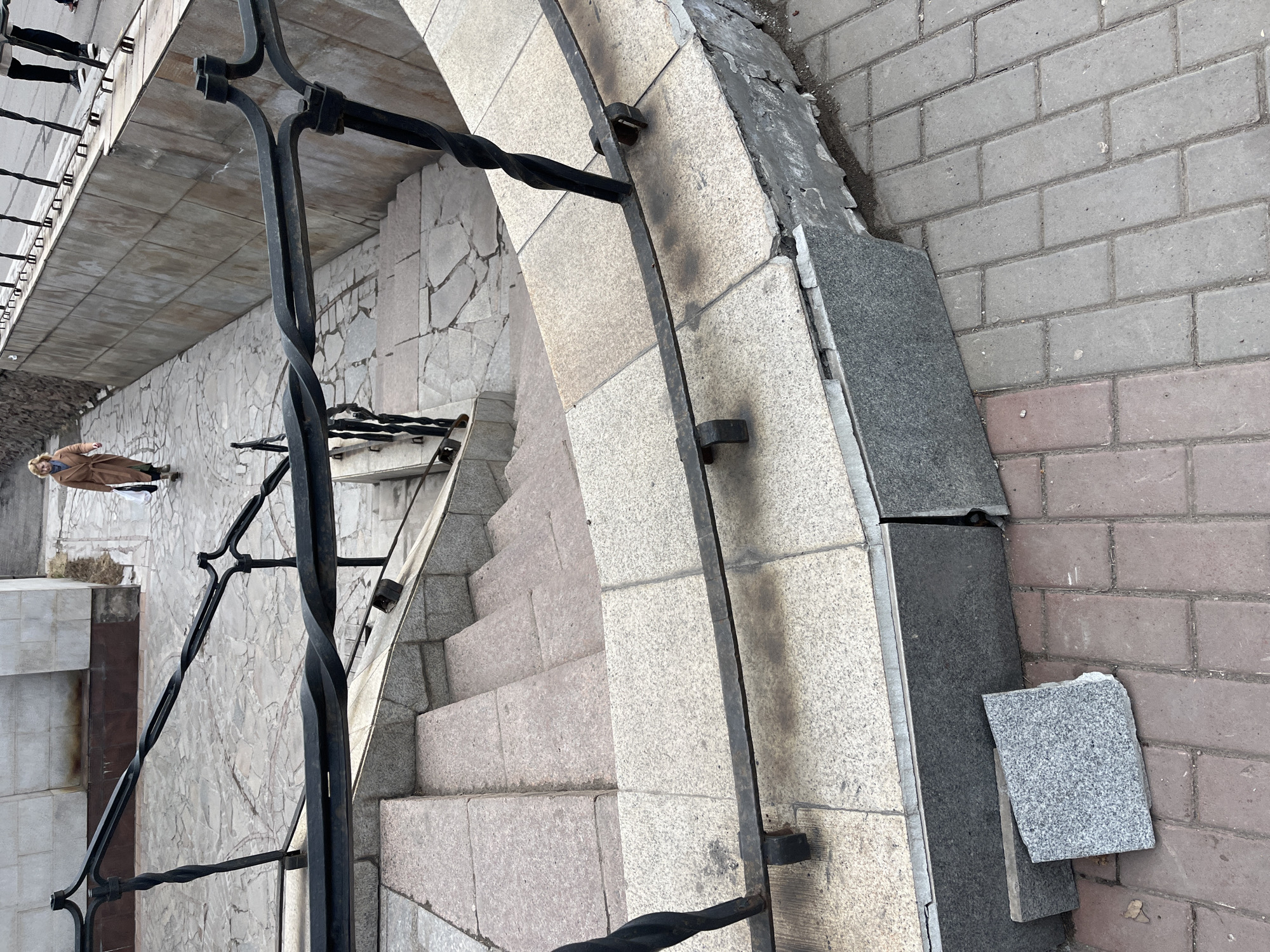 The image shows a stone staircase with a black wrought-iron railing, set in a brick-paved area in an urban environment, possibly a city. A person, wearing a long tan coat, is visible in the background walking on a stone-tiled surface near the staircase. Some architectural elements, such as stone walls, are also present in the surroundings. The location is Unknown, Unknown.