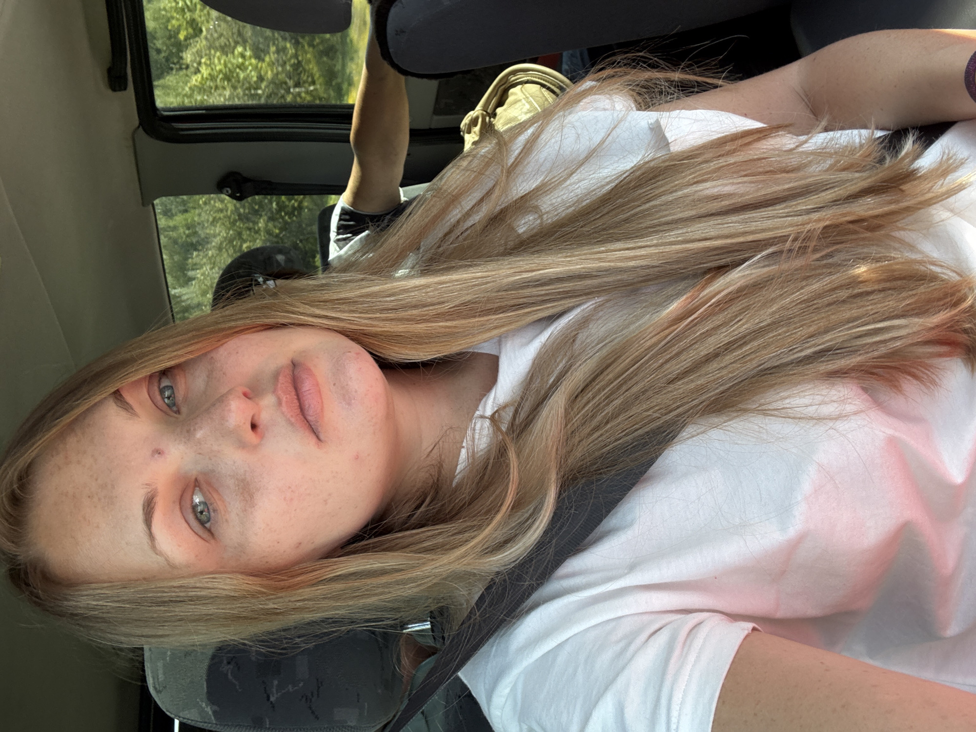 The image shows a close-up, selfie-style shot of a woman with long blonde hair and fair skin, freckles, and light blue eyes. She is wearing a white t-shirt and a seatbelt. The woman appears to be in the passenger seat of a vehicle, with a blurred background of trees and foliage visible through the window. Another person's arm is visible in the background, suggesting they are in the driver's seat. The lighting suggests it is daytime. There is no specific information to indicate the location is the city of "Меркурий, Russia".