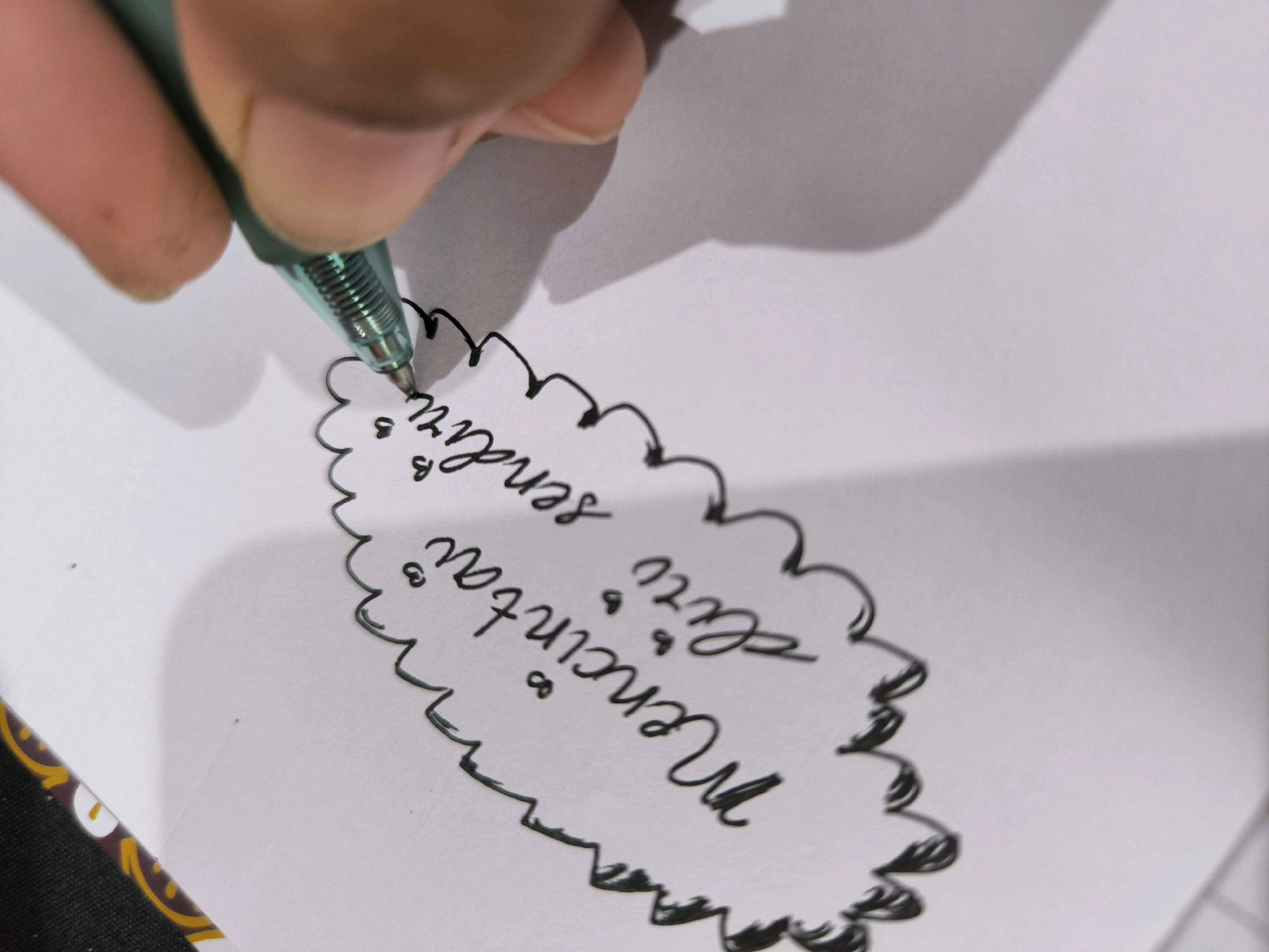 A close-up, high-angle shot captures a person's hand holding a teal gel pen, writing on a white piece of paper. The pen tip is actively making contact with the paper, creating a hand-drawn, cloud-like outline. Inside this outline, handwritten text is visible. The writing appears to be in Indonesian and says "Mencintai kamu selalu" which translates to "Loving you always." The handwriting is cursive and somewhat stylized. The background is a plain white surface, with a subtle shadow cast by the hand and pen. A hint of a patterned edge, possibly from a notebook or binder, is visible at the bottom left of the frame. The scene does not provide any specific geographic indicators to place it within Tabanan, Indonesia, or any other city.