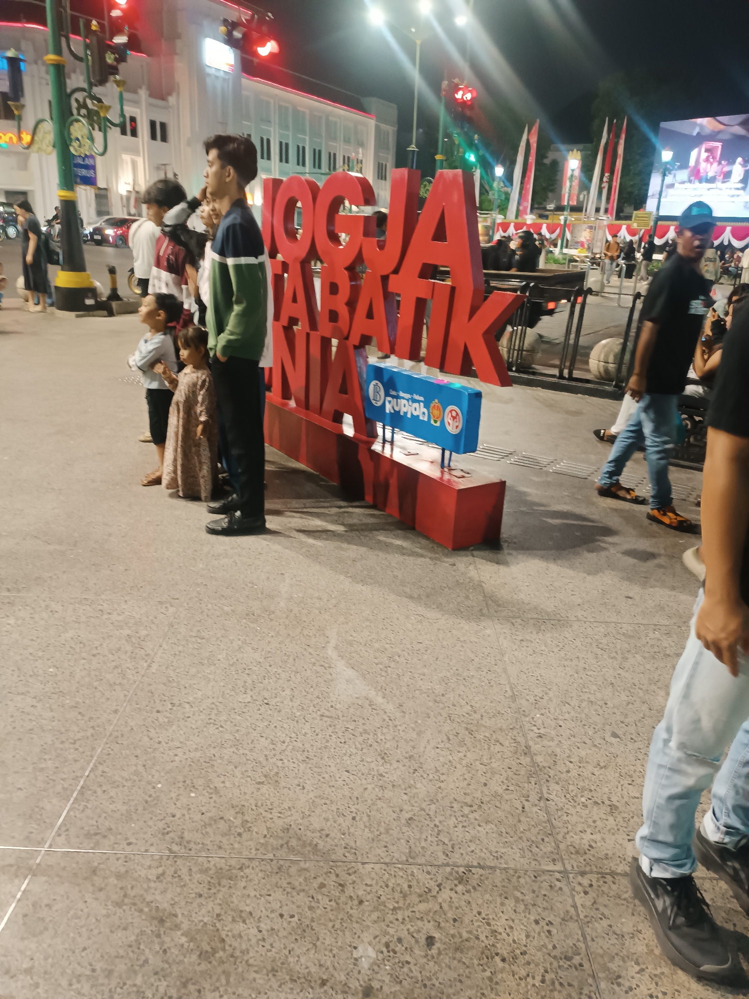This is a nighttime, eye-level, medium shot of a street scene in Yogyakarta, Indonesia. In the foreground, a large, red, three-dimensional sign reads "JOGJA BATIK". A blue sign with the word "Rupiah" and several icons is attached to the base of the "JOGJA BATIK" sign. A group of people, including several children and adults, are standing in front of the sign, facing left. They appear to be posing for a photograph or watching something in the distance. The street pavement is visible in the foreground, with metal grates embedded in it. In the background, there are buildings with red and white striped awnings, streetlights, traffic lights, and Indonesian flags. Several people are walking in the background. The overall atmosphere is festive and lively.