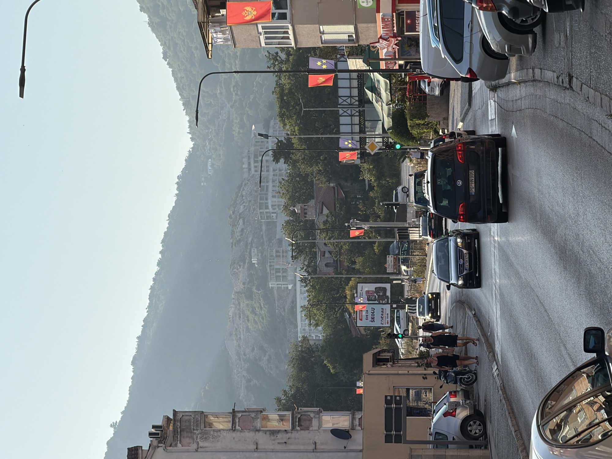 In Budva, Montenegro, on a sunny day, a two-lane road is lined with buildings and dense greenery on a hillside. Cars, including a silver hatchback, black Volkswagen Golf, and a black sedan, navigate the asphalt road. Pedestrians, two women in black dresses and sandals, walk on the sidewalk. The scene is illuminated by sunlight filtering through a light haze, casting shadows on the road. The buildings exhibit a mix of architectural styles, with some featuring balconies and decorative elements. Flags, likely national symbols, are displayed on poles. The mountainous landscape in the background hints at the city's coastal geography. A traffic light shows green, indicating clear passage for vehicles.
