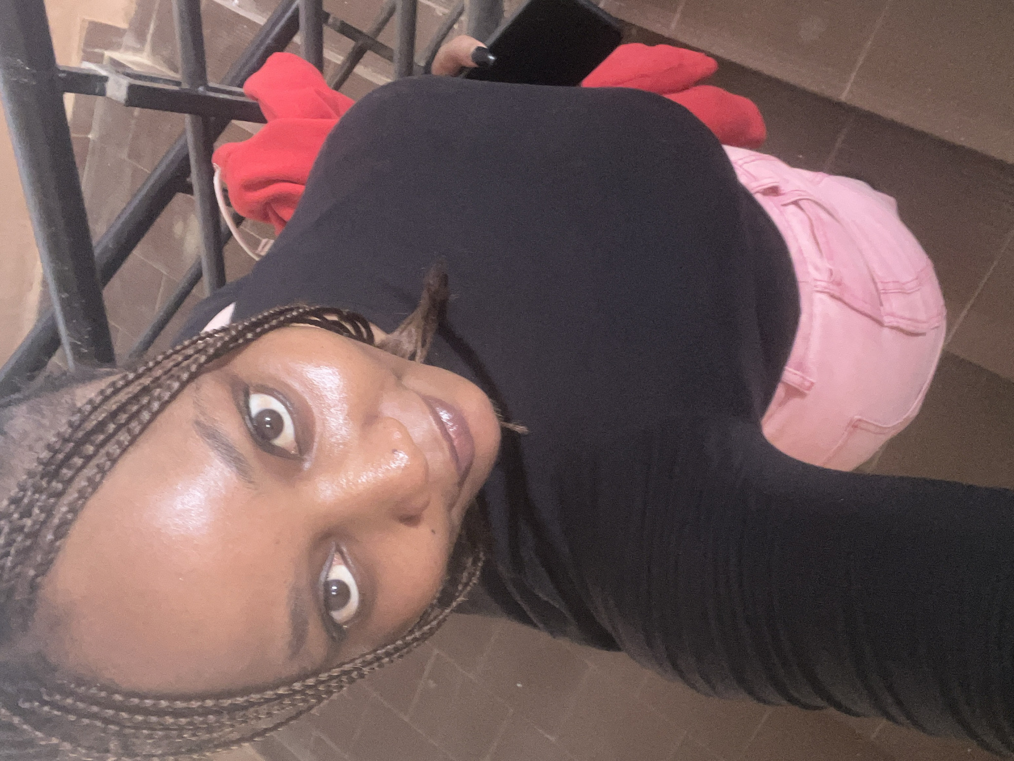 A high-angle, medium shot shows a Black woman with braided hair holding a black smartphone. She is wearing a black long-sleeved shirt and pink jeans. The background consists of a metal railing, reddish-brown steps, and a red garment draped over the railing. The scene appears to be a staircase, potentially in an indoor or covered outdoor area within the city of Jos, Nigeria.